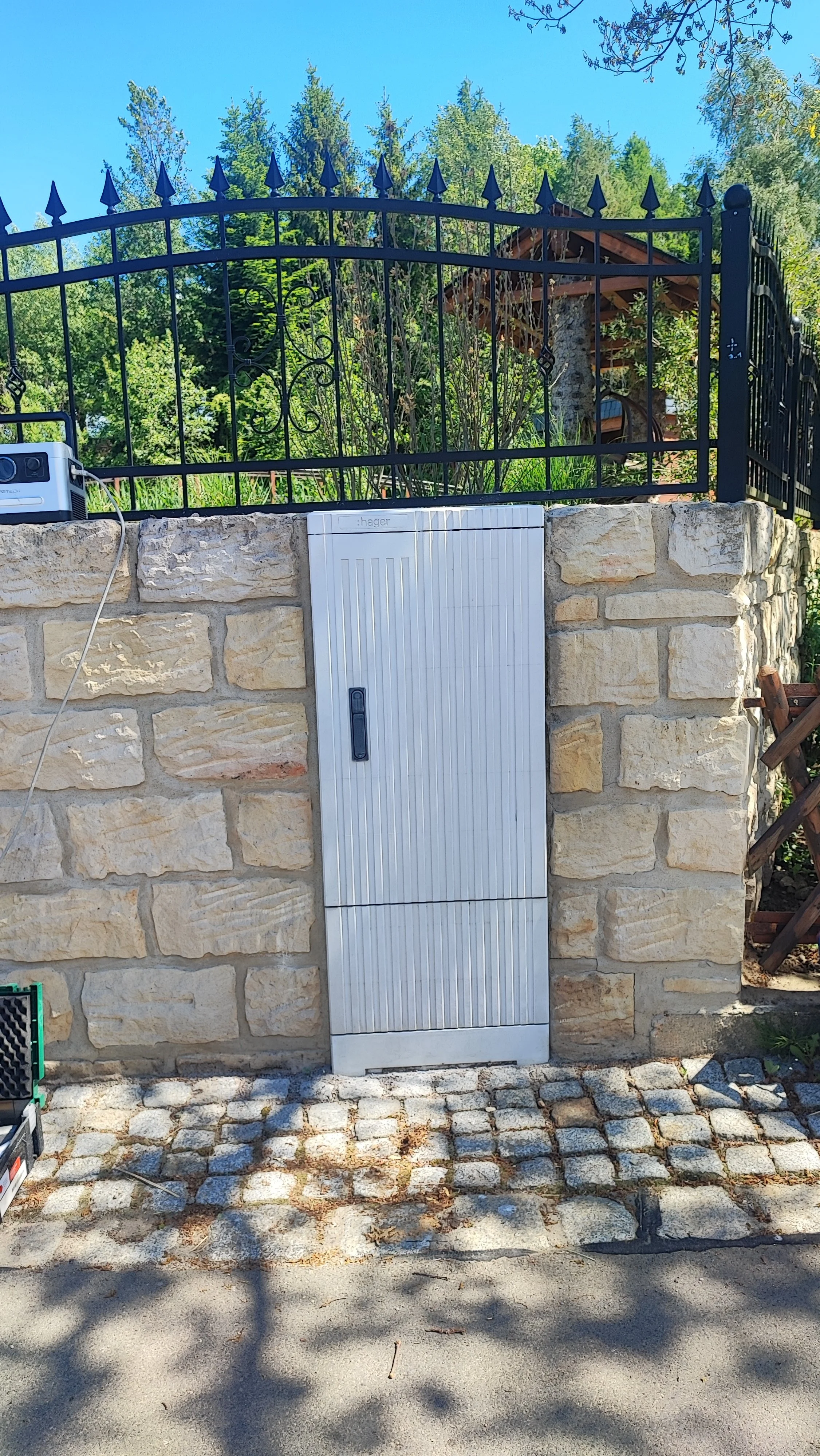 Ein Steinmauer mit einem metallenen Schaltkasten, darüber ein schwarzer Metallzaun und im Hintergrund Bäume und ein kleines Holzhaus.