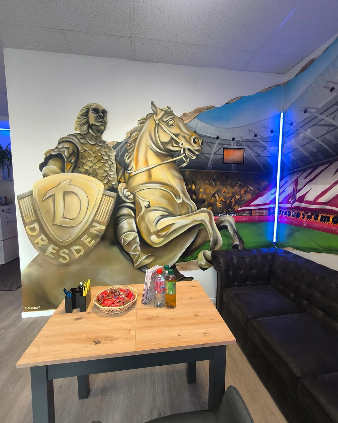 Wandbild eines Ritters mit Schild, einem Pferd und einem Stadtwappen, in einer modernen Einrichtung mit Tisch, Getränken und Sofa.