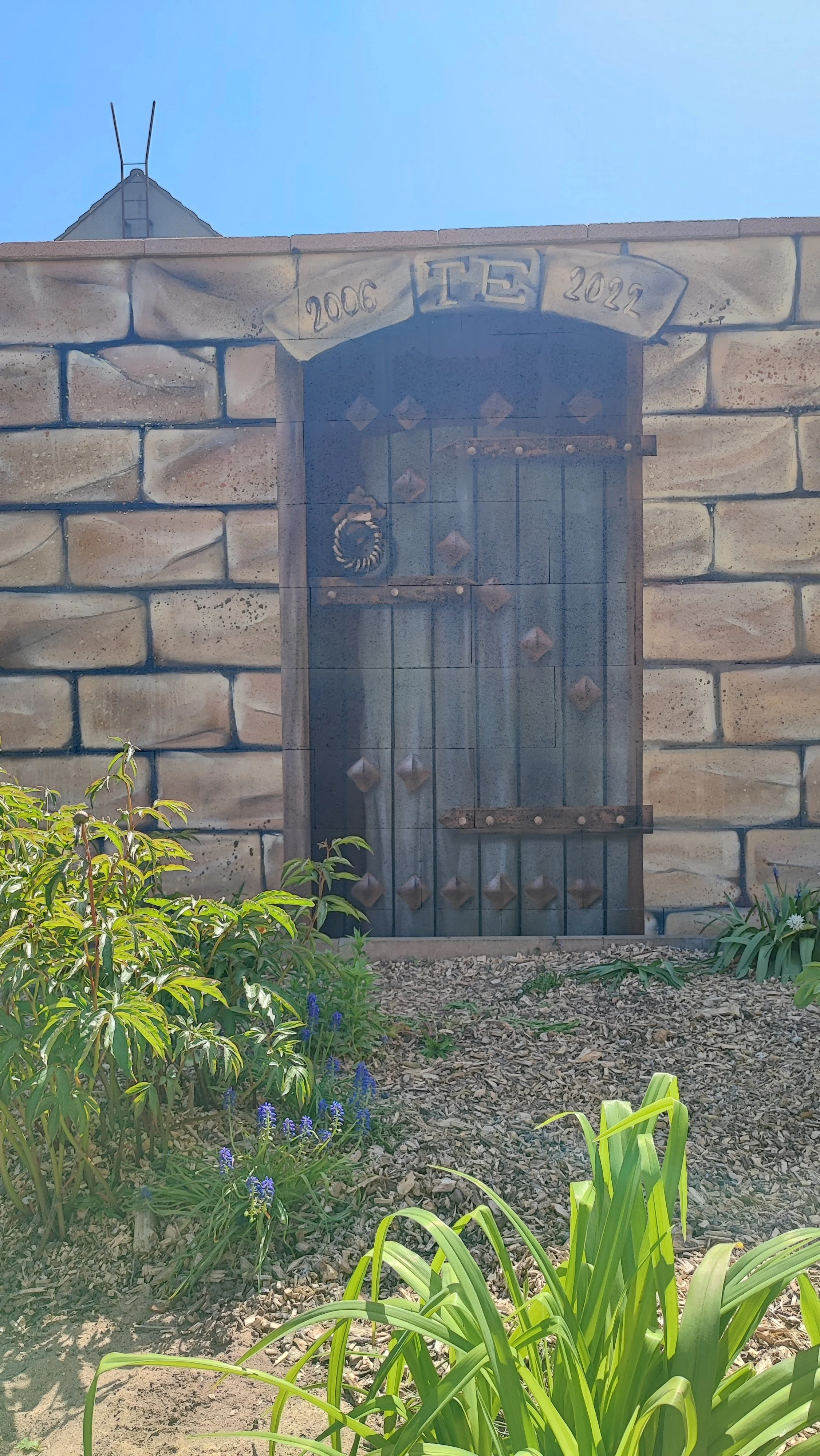 Holztür in einer Mauer mit einem Segel und dem Jahr 2006 sowie 2022 darüber. Im Vordergrund Blumen und grüne Pflanzen. 
