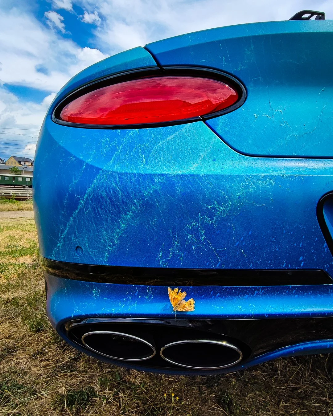 Der hintere Teil eines blauen Autos mit Kratzern, einem kleinen gelben Schmetterling auf der Stoßstange und einem Himmel mit Wolken im Hintergrund.