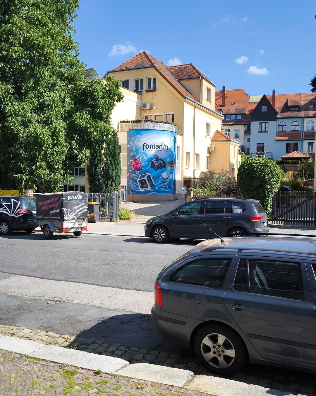 Straßenansicht mit geparkten Autos, bunter Wohnhäuser im Hintergrund, Baum und eine große Werbetafel mit dem Schriftzug "Fontaum" an einer hohen Wand.