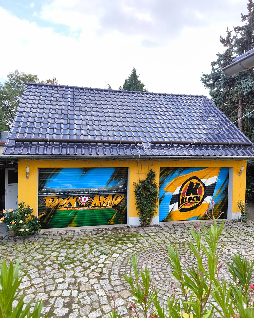 Ein gelbes Gebäude mit zwei Rolltoren, die mit Graffiti bemalt sind. Der rechte Gabentor hat das Logo "K-Block" und der linke das Wort "Dynamo" in Graffiti-Style. Das Gebäude ist von einer Steinpflasterstraße umgeben mit einigen Pflanzen davor.