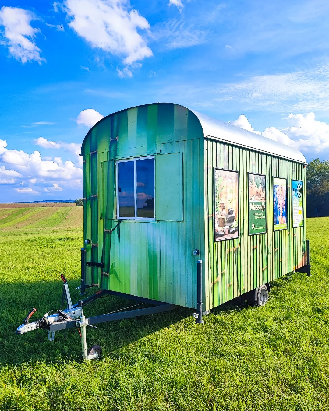 Grüner, kleiner Verkaufswagen mit Werbung für Massage-Dienstleistungen, steht auf einer grünen Wiese unter einem blauen Himmel mit Wolken.