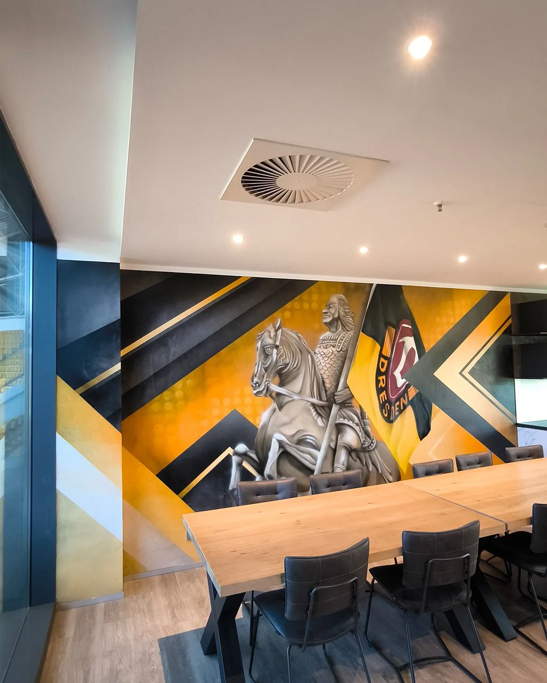 Abstrakte Wandmalerei mit einer römischen Kriegerstatue auf einem schwarzen Pferd, einem schwarzen und roten Flagge mit dem Text 'DRESDEN' und geometrischen Mustern in Schwarz, Gelb und Weiß, in einem modernen Sitzungssaal mit Holztisch und schwarzen