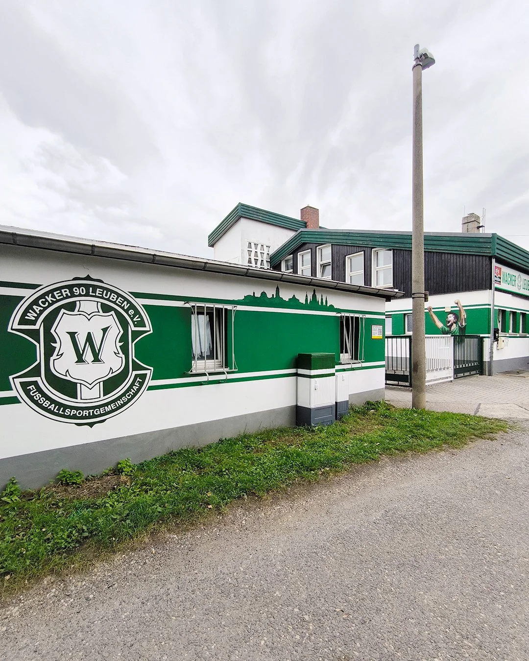 Außenansicht eines Fußballstadions mit Vereinslogo 'Wacker 90 Leuben e.V. Fußballgemeinschaft' an der Seite eines Gebäudes, darunter ein Wandbild eines jubelnden Fußballspielers