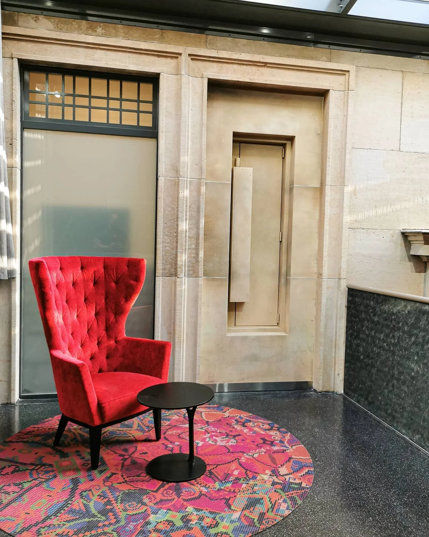 Ein roter, gepolsterter Sessel mit hoher Rückenlehne steht auf einem bunten, geometrischen Teppich. Im Hintergrund sind eine Tür und ein Fenster in einer beige Steinwand sichtbar.