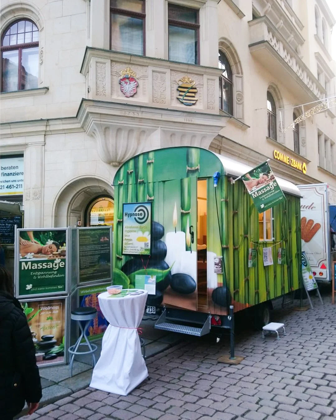 Mobiler Wellness- und Massageservice mit grün gestaltetem Trailer auf einer Stadtstraße, mit Informationsschildern und einem kleinen Stehtisch vor dem Trailer.