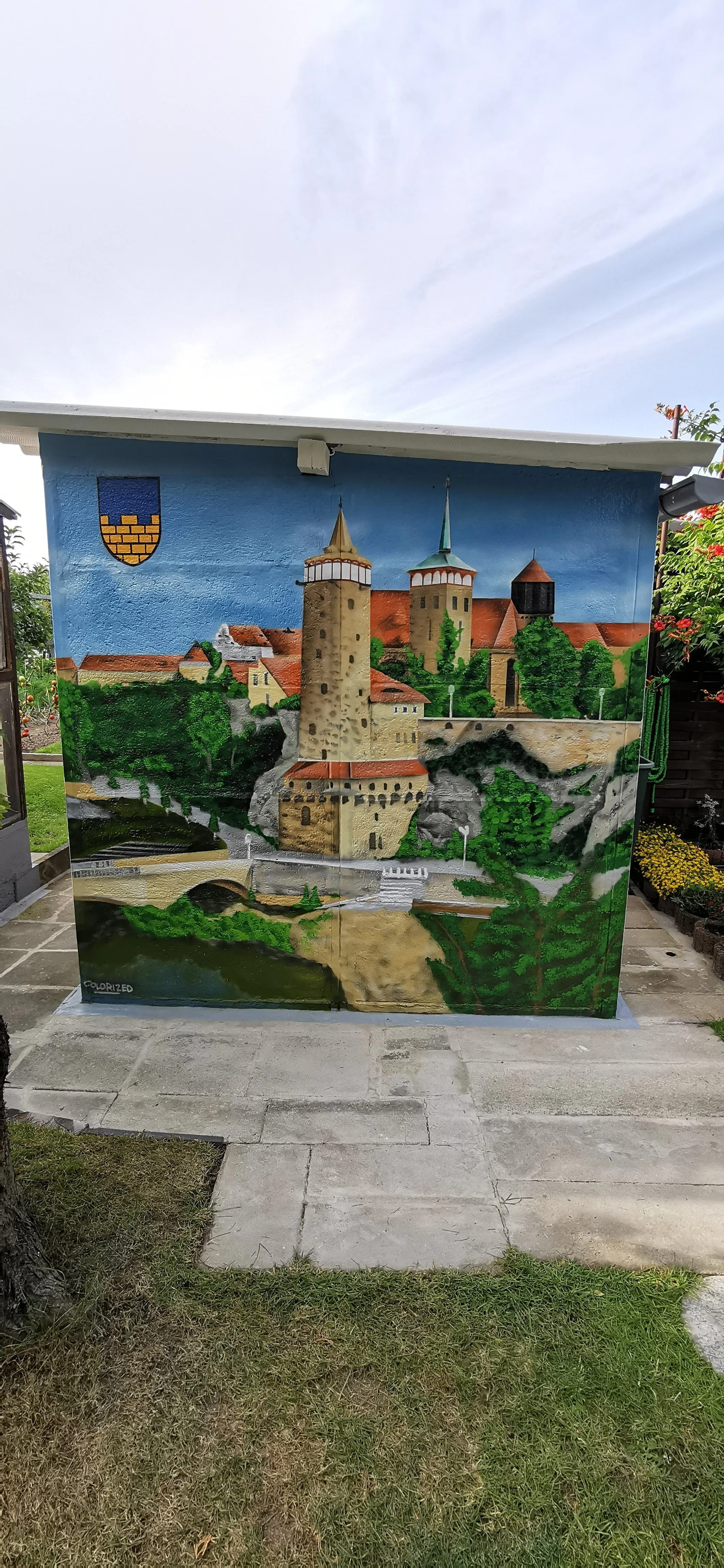 Eine bemalte Wand mit einer Darstellung eines mittelalterlichen Schlosses auf einer kleinen Struktur im Garten. Das Gemälde zeigt Türme, Mauern, Bäume und einen Fluss.