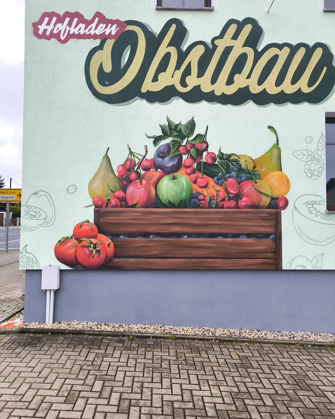 Grafik einer Obstkiste mit diversen Früchten wie Äpfeln, Birnen, Trauben und Tomaten auf einer hellblauen Wand. Über der Obstkiste steht der Schriftzug 'Hofladen Obstbau'.