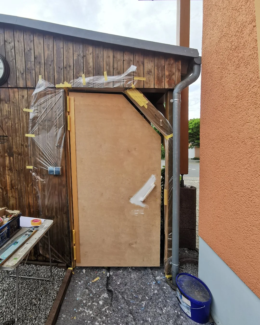 Ein Holzschrank wird an einer Außenwand montiert, eingerahmt von einer Holzfassade und einer orangefarbenen Wand. Es gibt Werkzeuge und Malerband auf einem Tisch daneben.