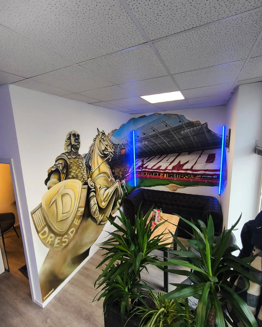 Eine Ecke in einem Wartebereich mit Wanddekoration, einem schwarzen Sofa, einem Tisch mit Getränken und grünen Pflanzen, sowie einem dekorativen Hintergrund mit Neonlicht.