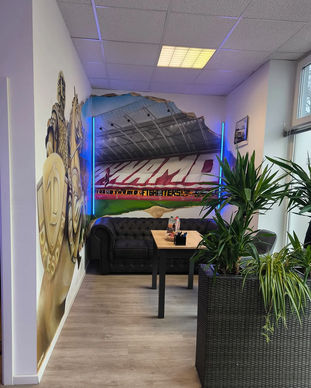 Ein Wartebereich mit dunklem Ledersofa, einem kleinen Tisch mit Schreibutensilien, einer großen Pflanze in einem Korb, einer Wandbemalung eines Fußballstadions und einem Sportlogo.