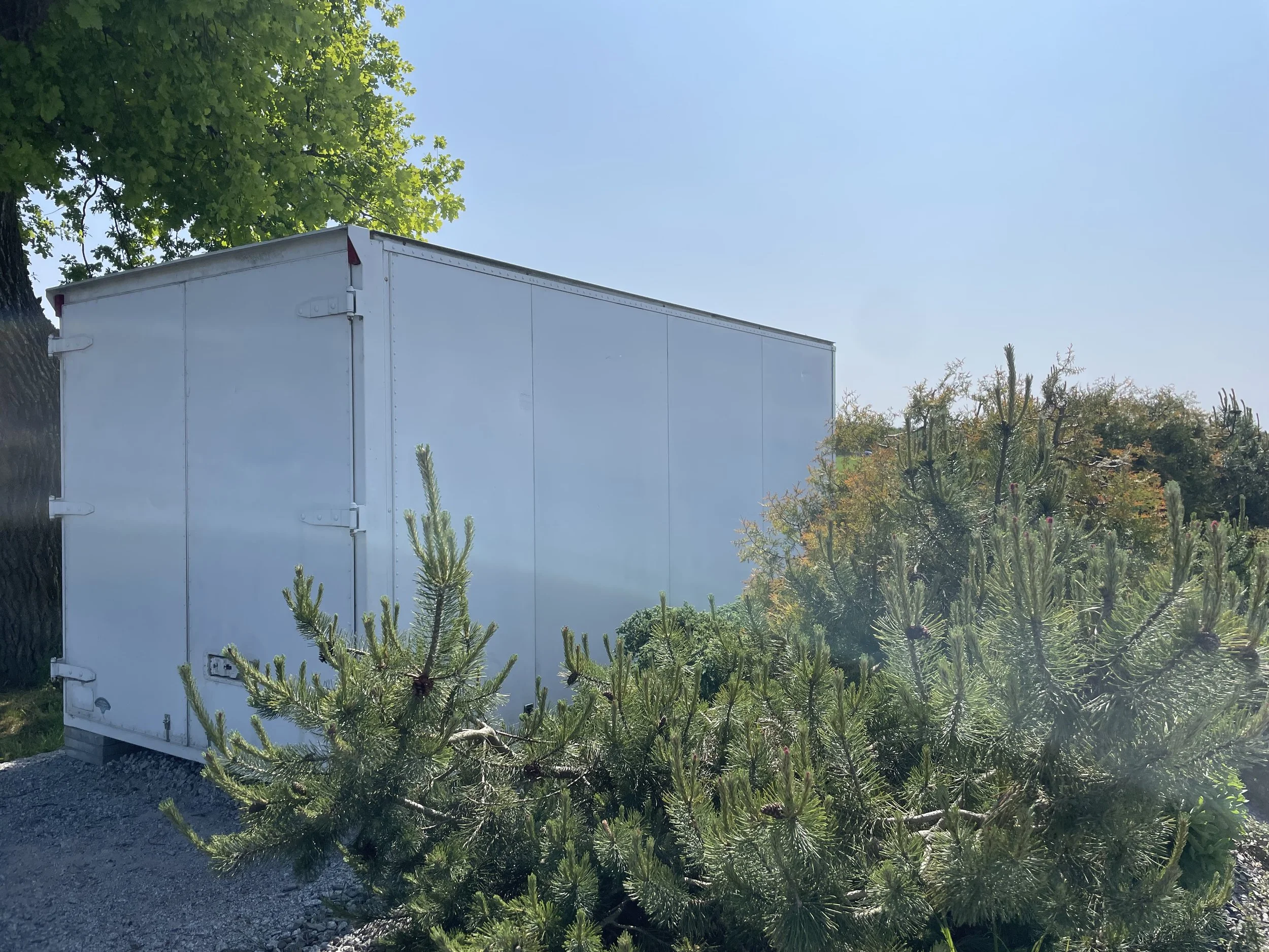 Weiße Lagerbox im Freien, umgeben von grünen Büschen und Bäumen bei sonnigem Himmel.