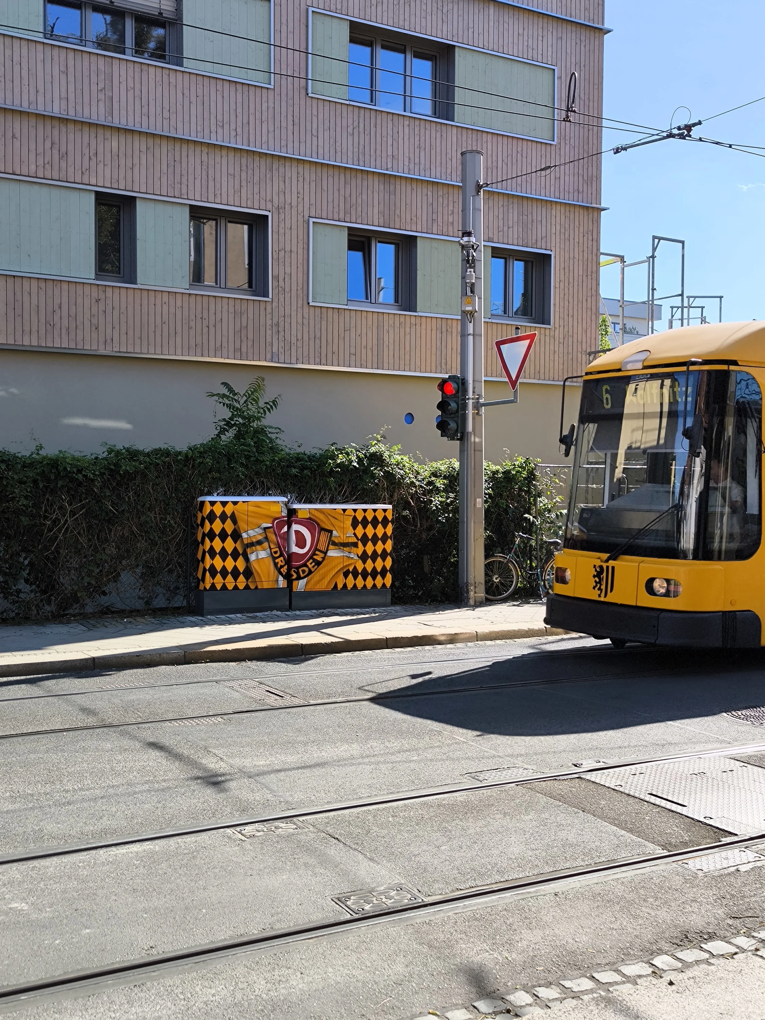 Eine Straßenkreuzung mit einem gelben Tram, einem Verkehrslicht und einem Abfalleimer mit dem Dresdner Eisteam, im Hintergrund ein modern gestaltetes Wohngebäude mit mehreren Fenstern.