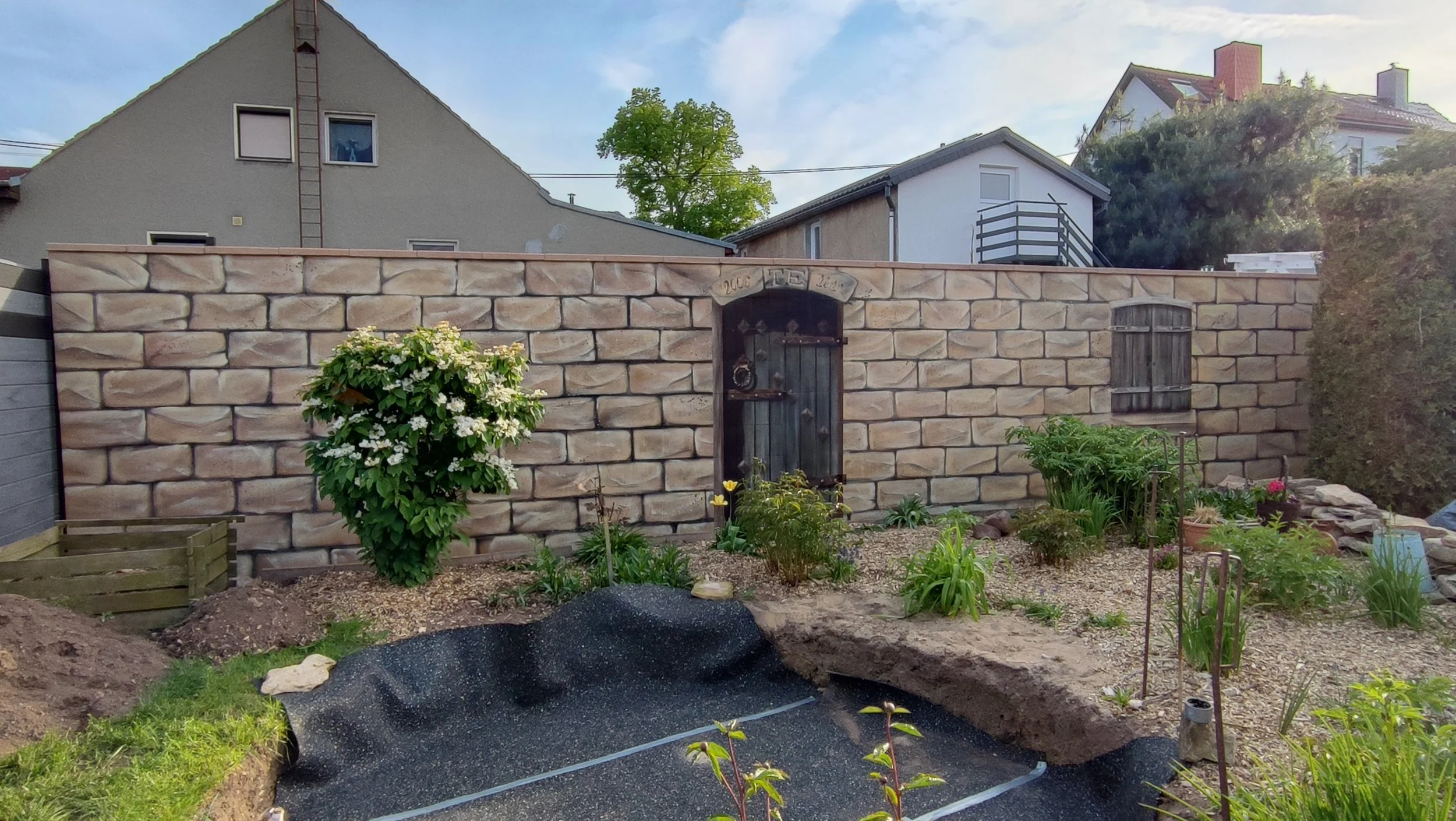 Garten mit Steinmauer, kleiner Tür, Blumentöpfe und Pflanzen, im Hintergrund Häuser und Bäume, sonniges Wetter.