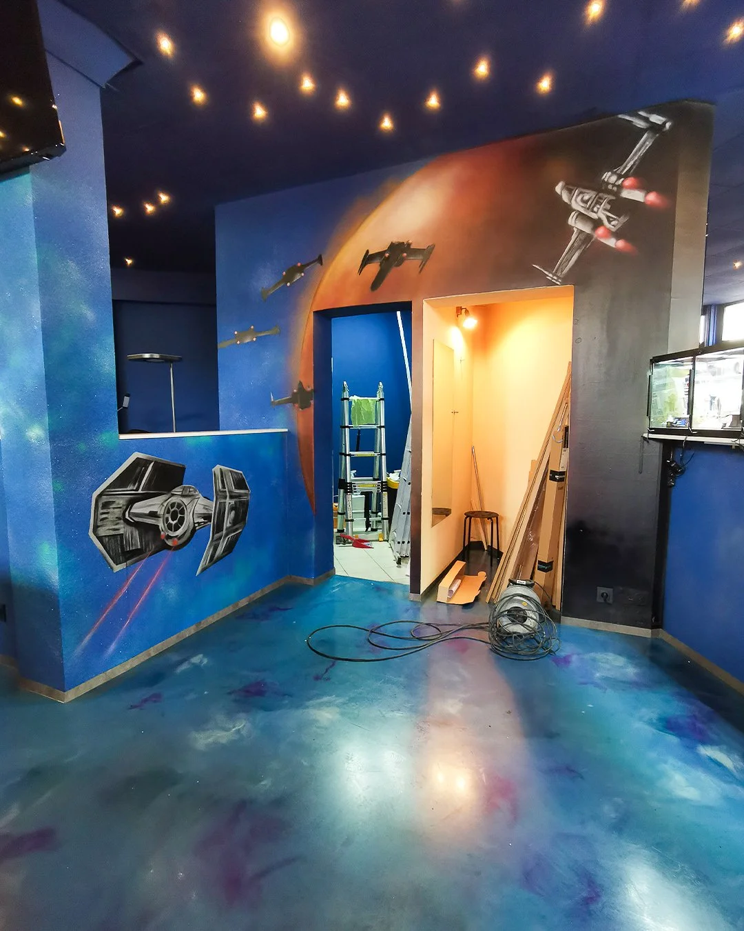 Innenraum mit Weltraum-Motiv, Wandmalerei zeigt Raumschiffe und eine Planetenoberfläche, Werkzeug und Leitungen im Raum, vermutlich eine Raumgestaltung oder ein Spielplatz mit Star Wars Thema.