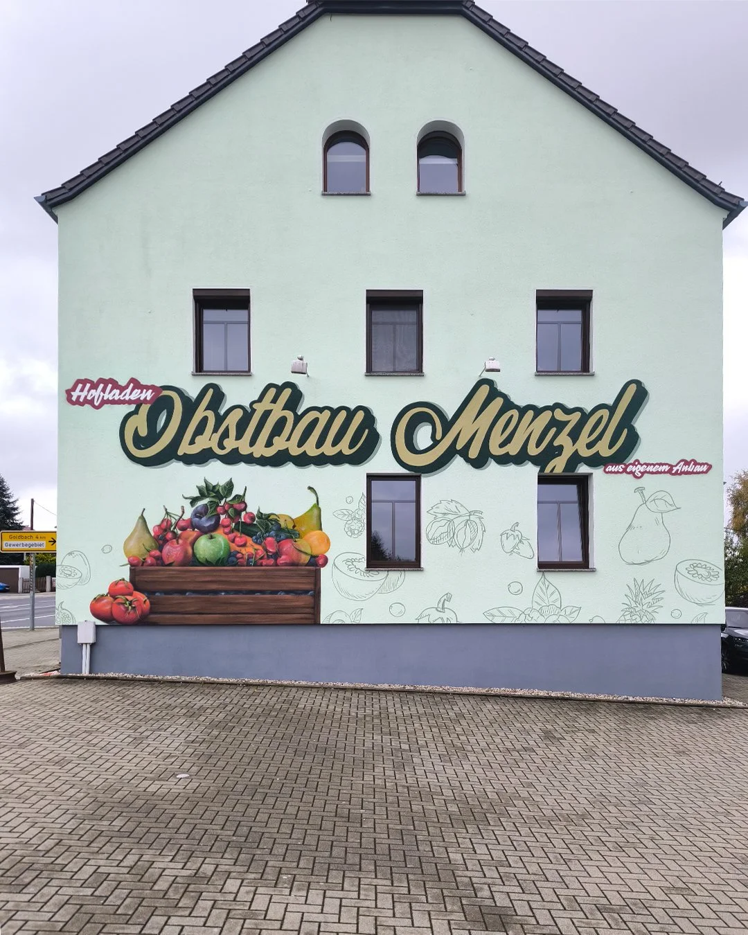 Fassade eines Gebäudes mit einem großen Wandbild, das einen Korb mit einer Vielzahl an Früchten zeigt, sowie den Text 'Hofladen Obstbau Menzel'.