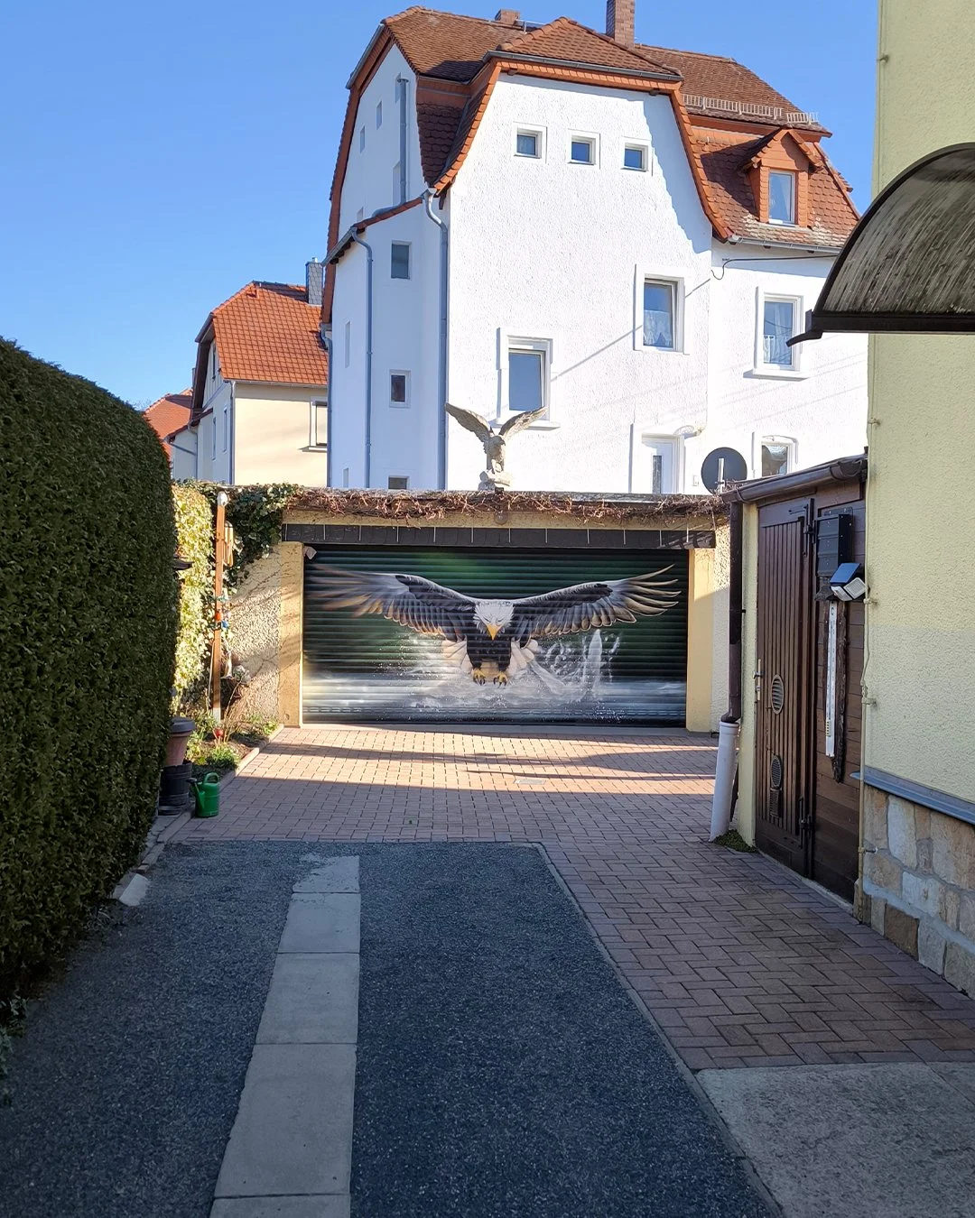 Gartentor mit Adler-Graffiti und sternförmigem Solarlicht, umgeben von Gebäuden und Hecken in einer sonnigen Straße.