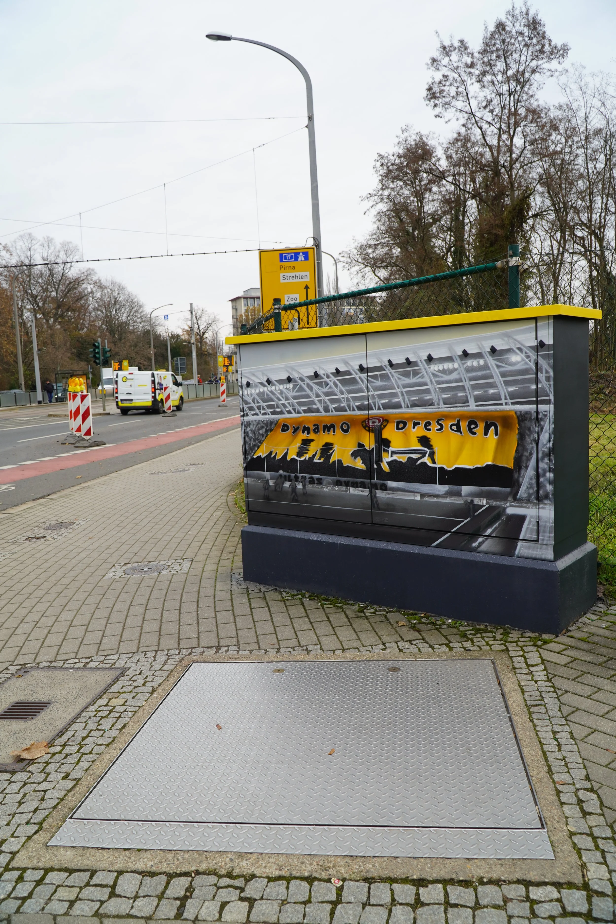 Stromkasten mit Dynamo Dresden Fan-Design an einem Gehweg in Dresden, in der Nähe einer Straße mit Fahrzeugen und Verkehrsampeln, im Hintergrund Bäume und eine gelbe Wegweiser-Tafel.