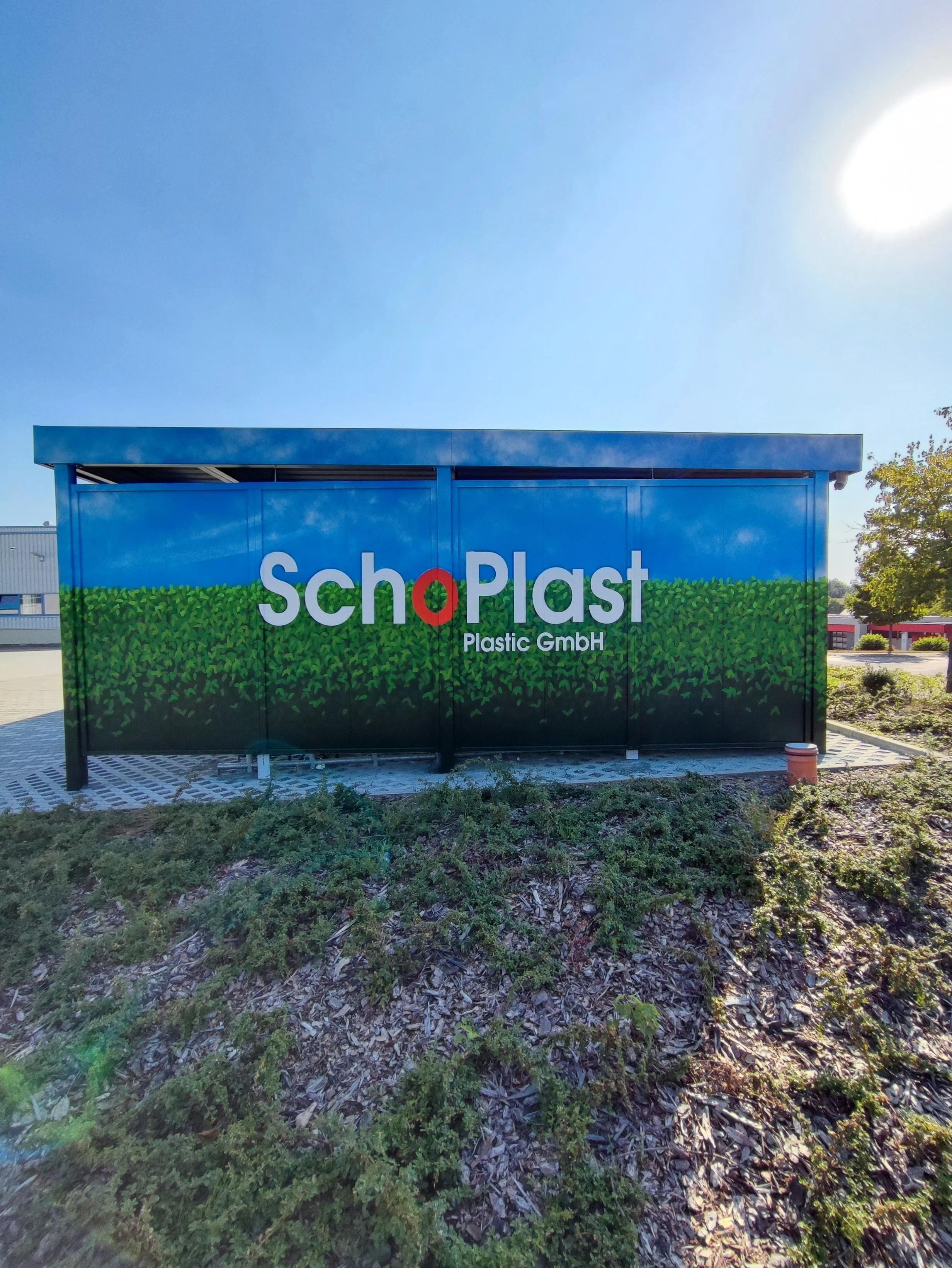 Werbeschild mit dem Schriftzug "SchoPlast" auf einem blauen Gebäude, darunter steht "Plastic GmbH", mit grüner Pflanzenmuster im Hintergrund und Sonnenlicht am Himmel. Graffiti Wandgestaltung in Dresden – Auftragsgraffiti, Workshops und kreative Graf