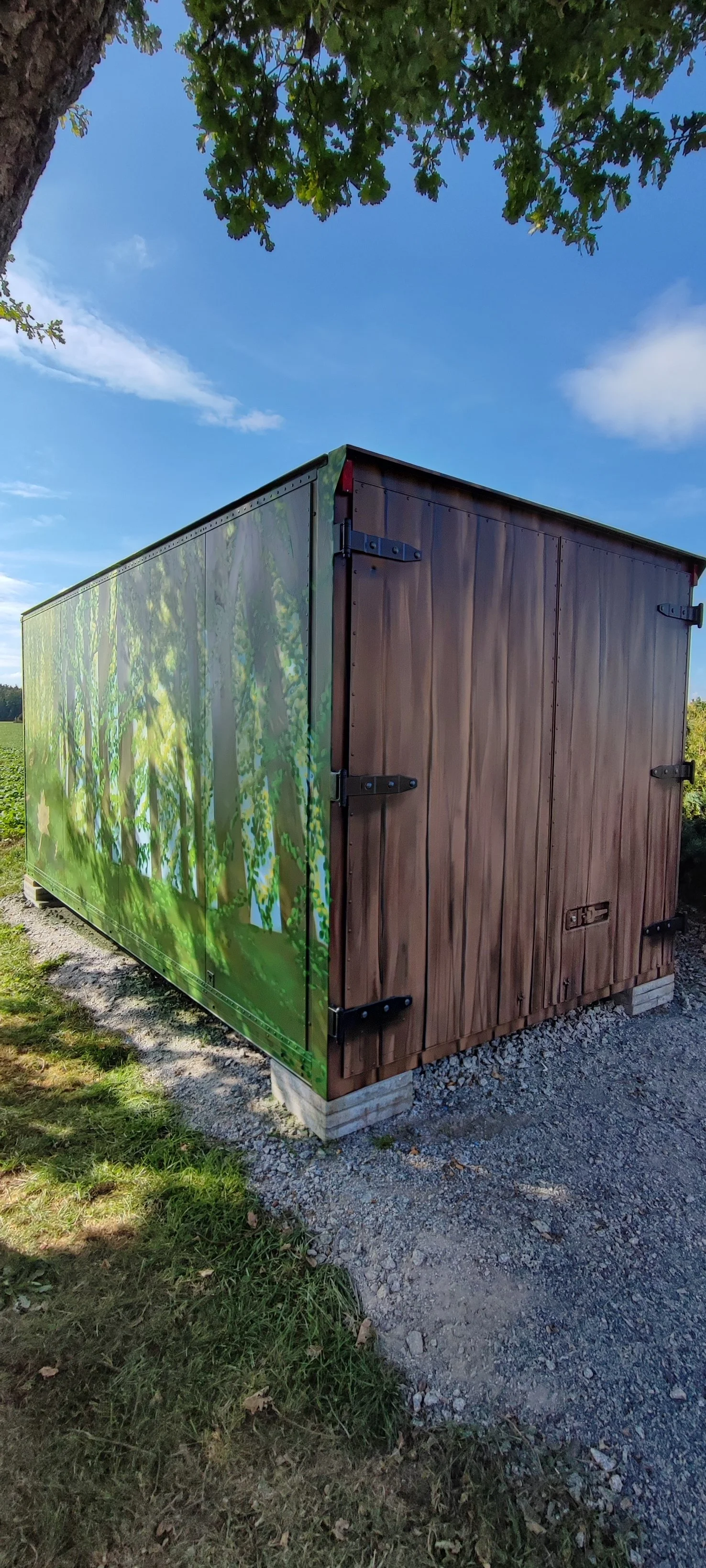 Ein Lagercontainer mit einer bunten Waldszene auf der Seite, steht auf einer Gras- und Kiesfläche, umgeben von Bäumen und einem blauen Himmel mit wenigen Wolken.