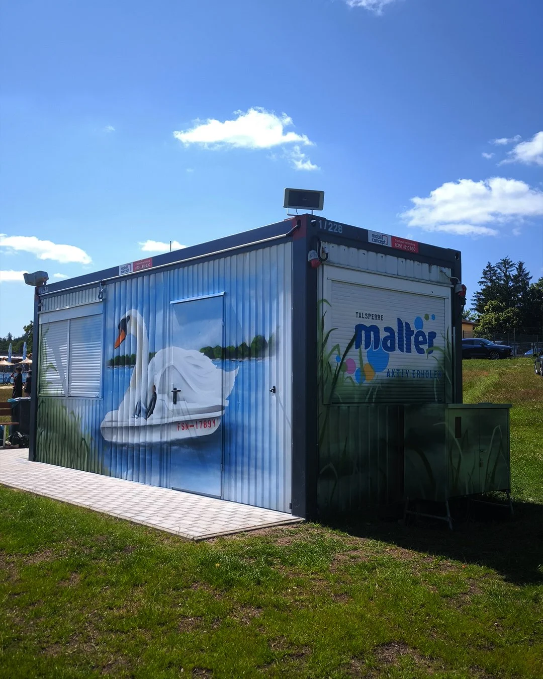 Kunstvoll bemalter Wassertank mit einem weißen Schwan auf einer Wasserfläche, umgeben von grünen Kiefern, mit dem Logo "maler" und der Aufschrift "Aktiv Erholen" auf der Seite. Professionelle Graffitigestaltung in Dresden: individuelle Wandgestaltung
