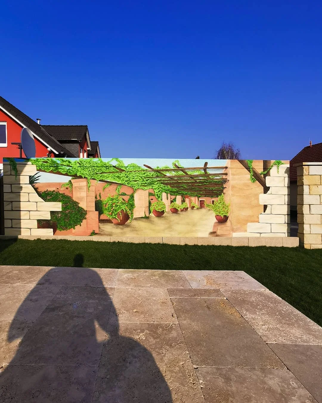 Ein Wandgemälde zeigt einen Garten mit hängenden Blumentöpfen, umgeben von Steinmauern, auf einer betonierten Terrasse vor einem blauen Himmel.