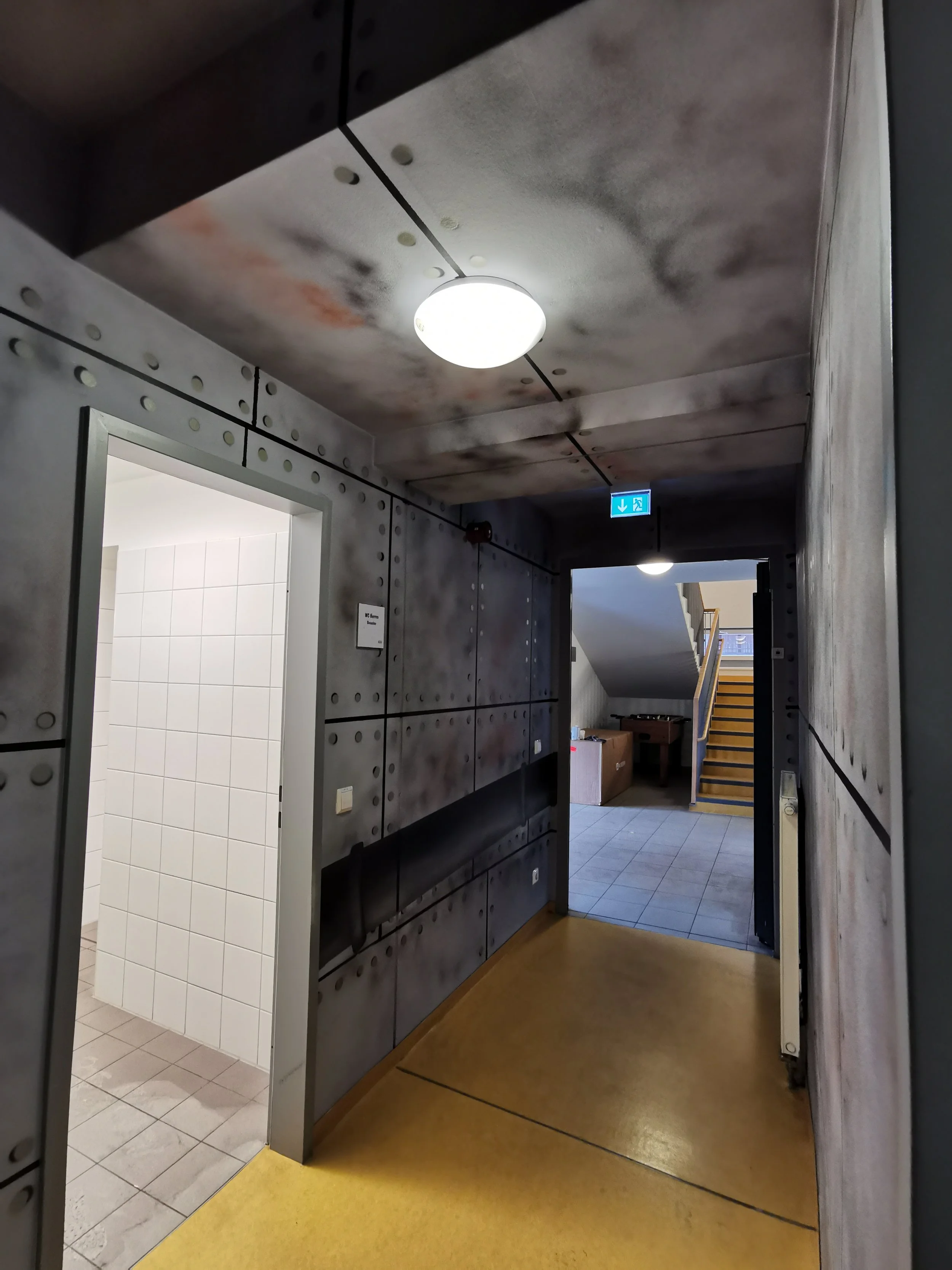 Ein Flur im Gebäude mit dunklen, metallen-ähnlichen Wänden und Decke, einer Deckenlampe, einem Durchgang zu einer Toilette auf der linken Seite, einem Ausgang mit Notausgangsschild nach oben, einem Treppenhaus im Hintergrund und einer gelben Bodenflä