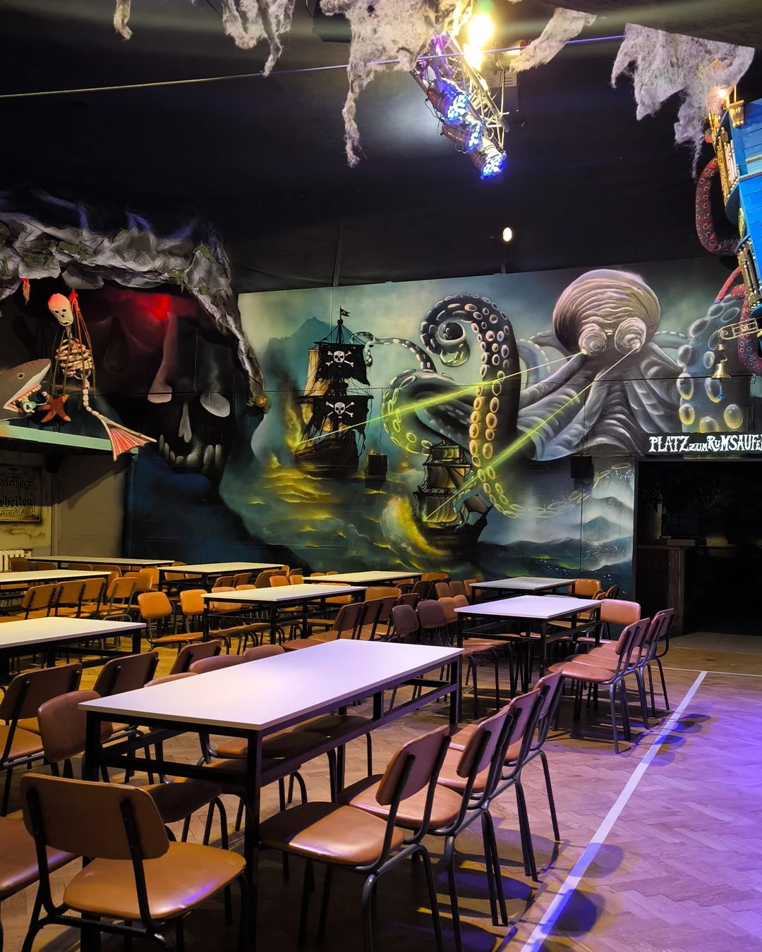 Eine Indoor-Szene mit mehreren Tischen und Stühlen, lebhafte Wandmalerei mit Piratenschiff, Kraken, Totenkopf und Deko-Elemente, die auf einer Halloween- oder Piratenausstellung hinweisen.