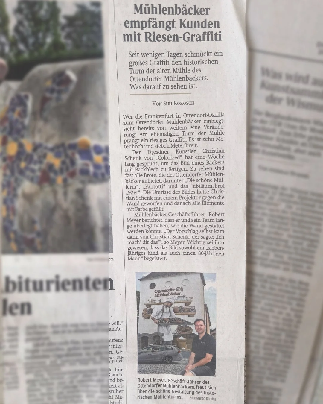 Zeitungsartikel mit Überschrift, dass die Mühlenbäcker Kunden mit Riesen-Graffiti empfangen. Es zeigt ein graues Wandbild eines Mühlenturms mit Bäckerei-Elementen, darunter eine Person, die vor dem Gemälde sitzt.