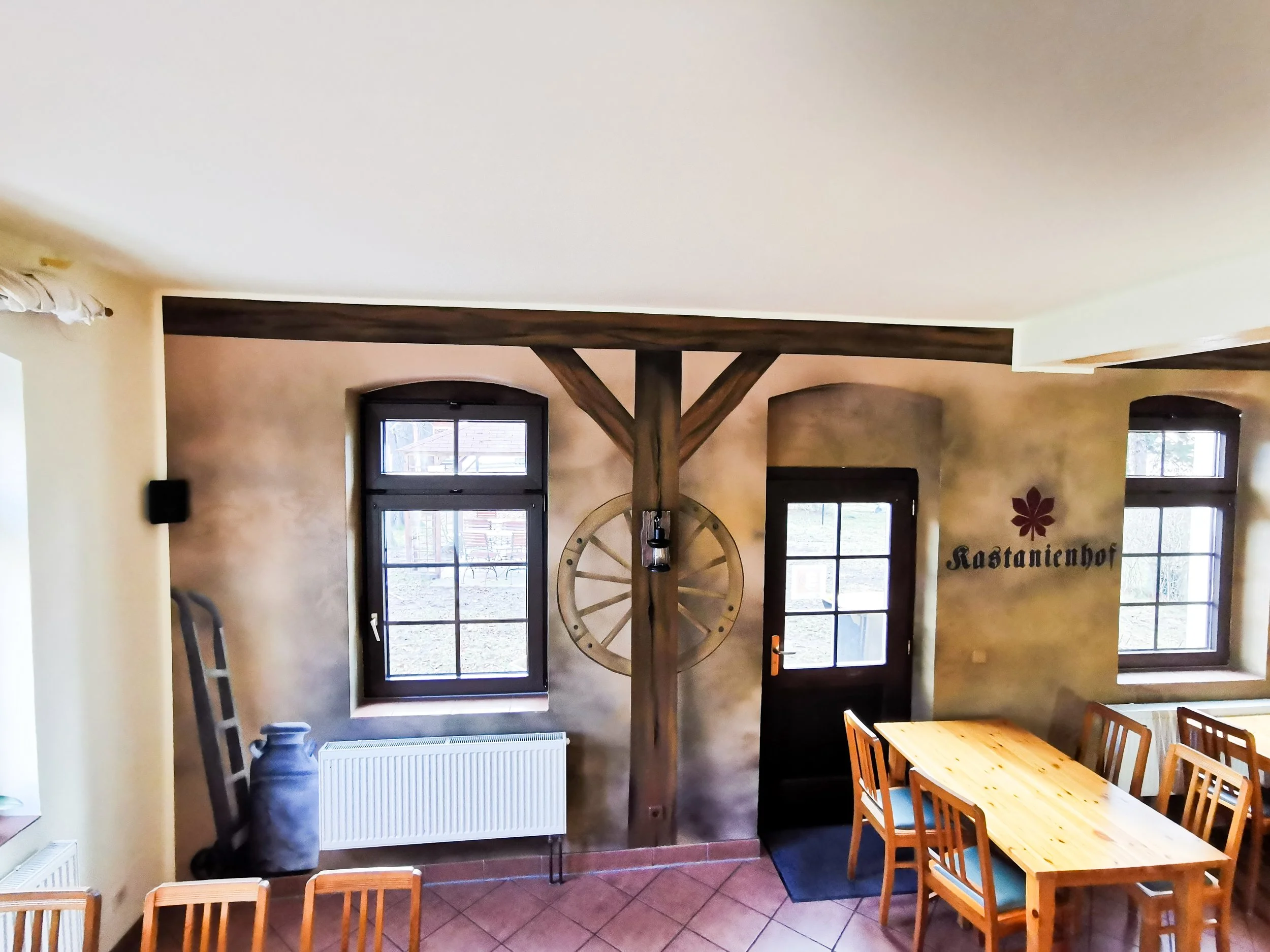 Innenraum eines rustikalen Restaurants mit Holztisch und Stühlen, Wanddekoration mit einem Holzwagenrad und einem Schriftzug 'Kastanienhof', zwei Fenster, ein Tür, und eine Wand mit warmen Erdtönen.