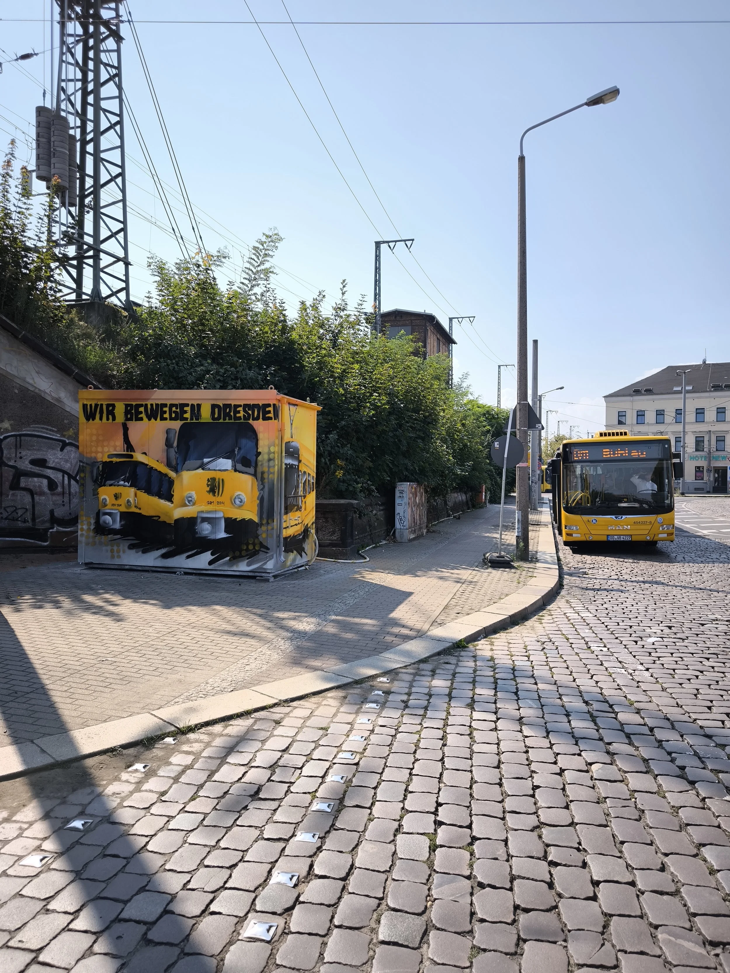 Straßenansicht in Dresden mit Bus und Graffiti an einem Container, farblosen Platten und Kopfsteinpflaster, grünem Gebüsch und Gebäuden im Hintergrund bei sonnigem Wetter.