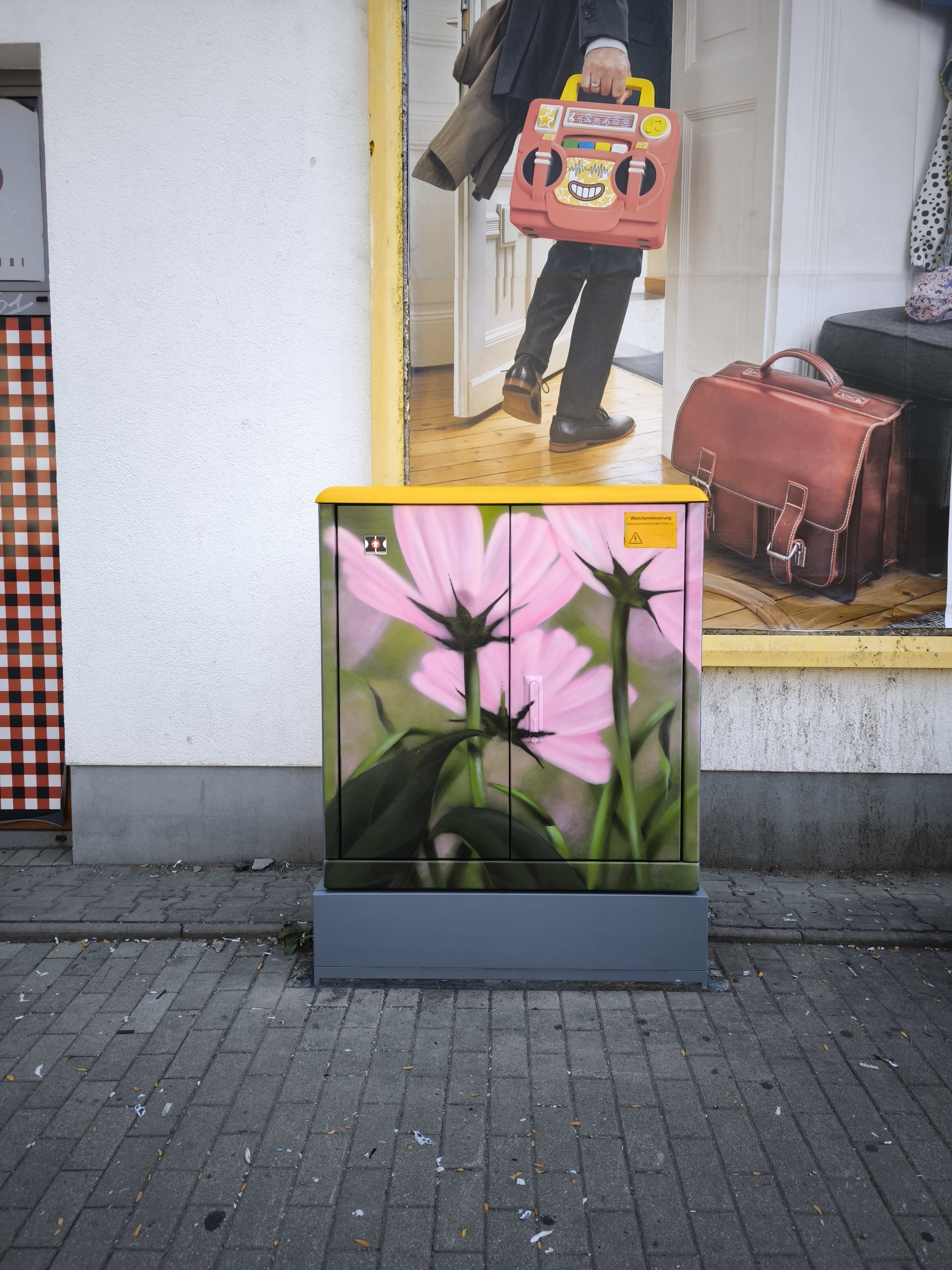 Auf dem Bild ist ein grauer Stromkasten mit einem überdimensionalen Bild von pinken Blumen zu sehen. Im Hintergrund befindet sich eine reflektierende Glasscheibe, die den Eingang eines Gebäudes mit einem Mann, der einen roten Boombox trägt, zeigt.