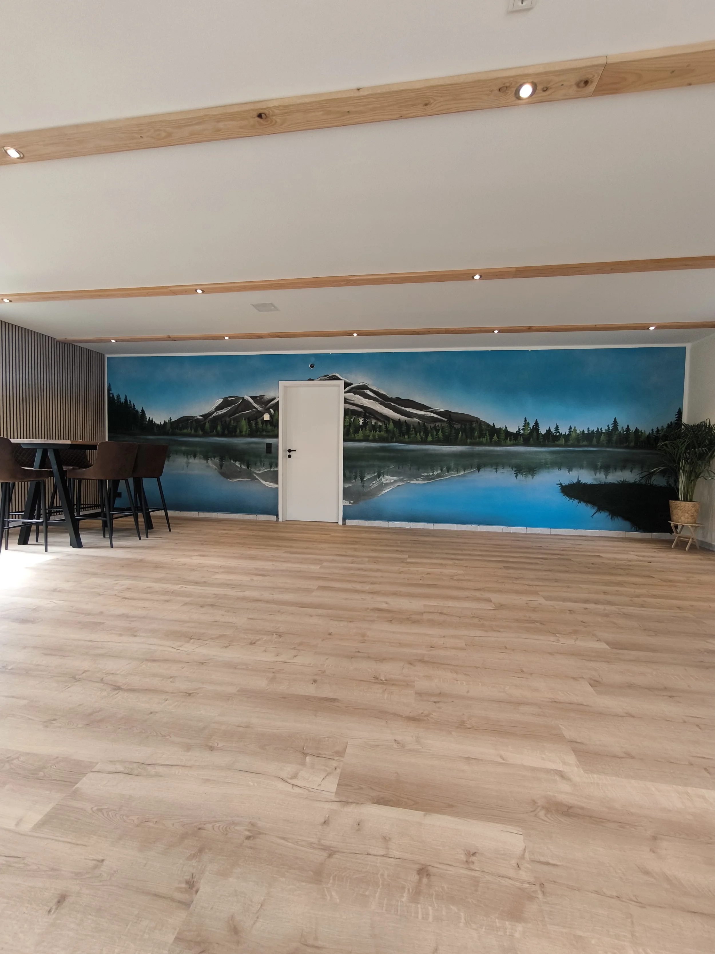 Ein Raum mit Holzfußboden, einer weißen Tür, einer Wandmalerei eines Sees mit Bergen und Bäumen, und einer Sitzgruppe mit hohen Stühlen.