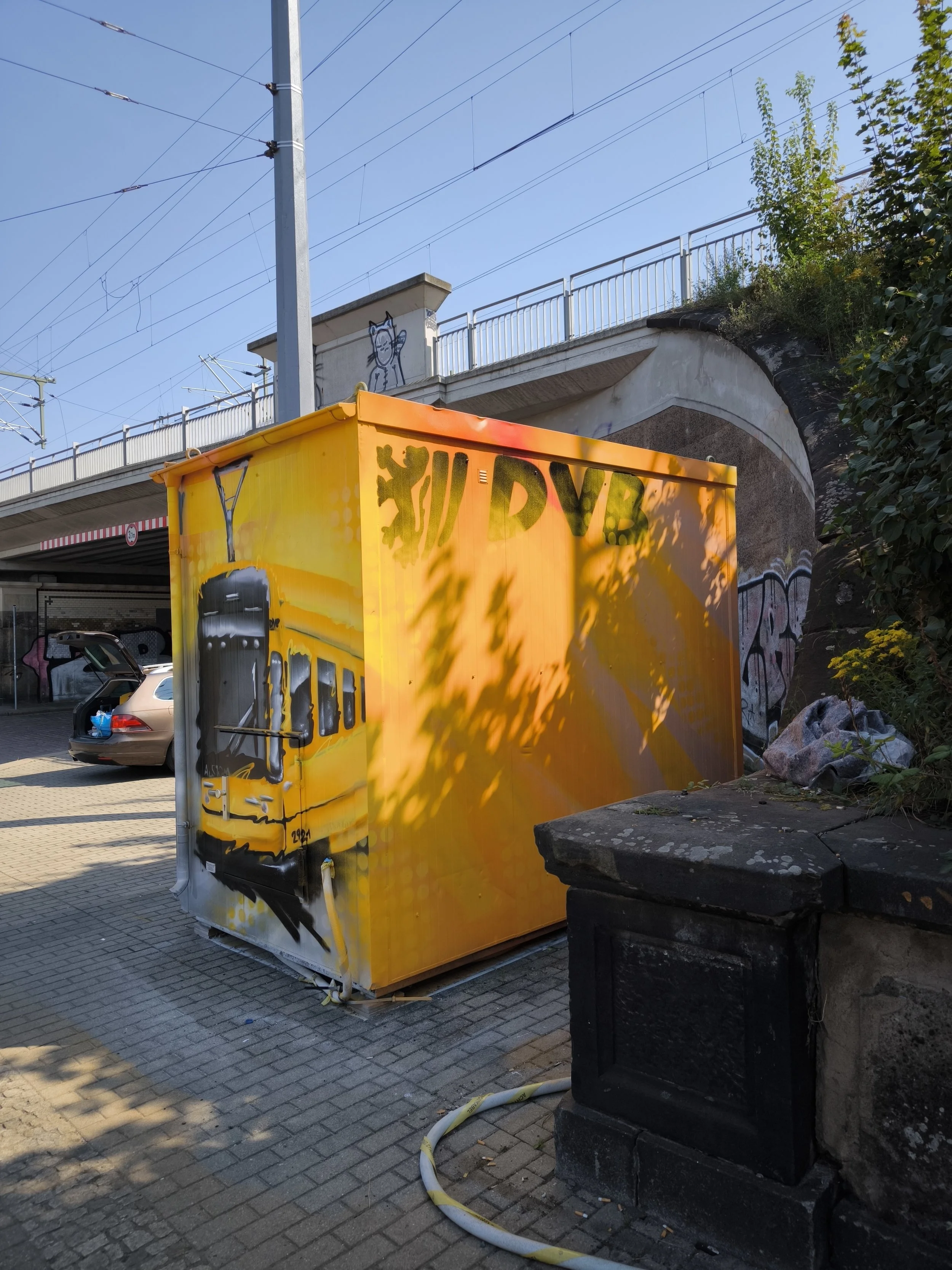 Graffiti-Kunstwerk eines gelben Zuges auf einem kleinen Kasten, umgeben von Schatten und Bäumen, im Hintergrund eine Brücke mit Wänden und Straßenbahnlinien.