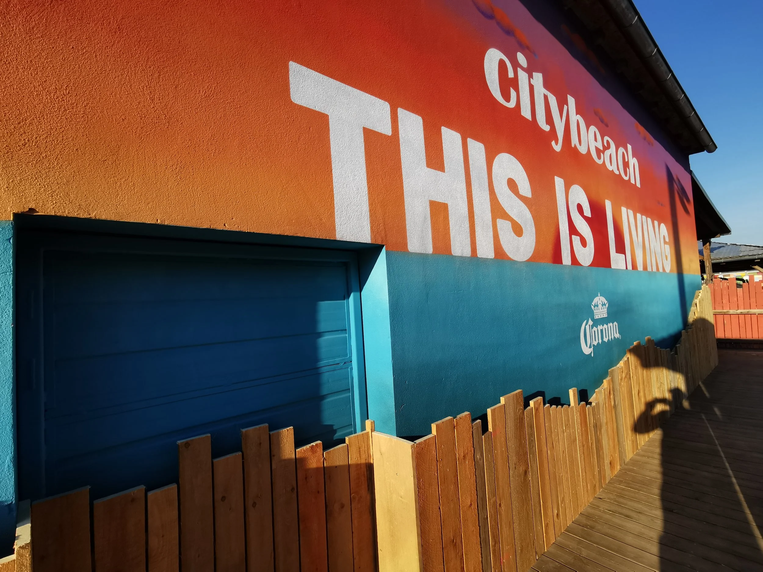Bunt bemalte Wand mit billboard-Text, Schriftzug 'citybeach', 'THIS IS LIVING', und 'Corona', umgeben von Holzpfosten und mit Holzdeck.