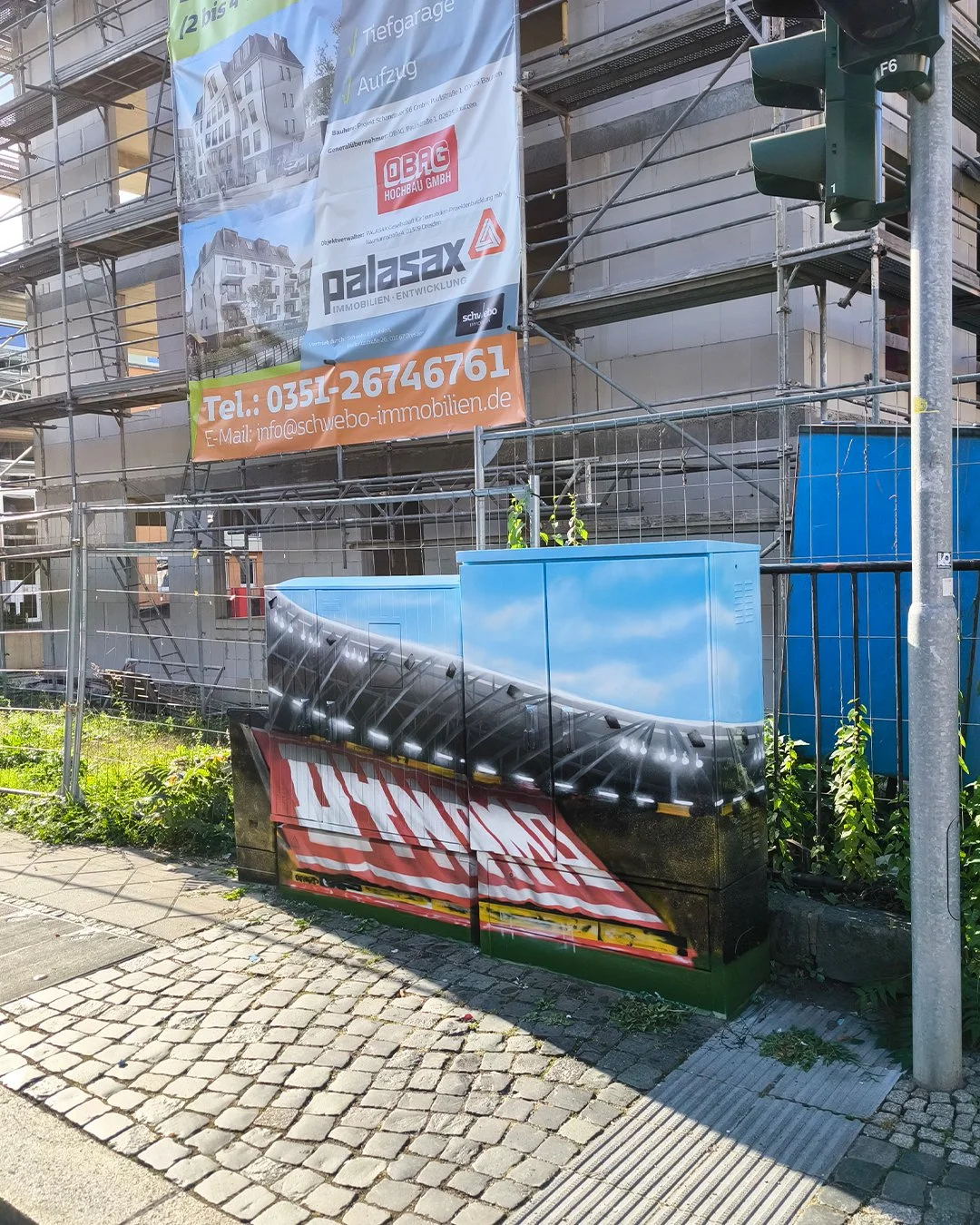 Baugerüst mit Bauzaun, Aufkleber mit Wolken- und Himmelmotiv, darunter eine Fotografierung eines Gebäudes und ein großes Banner mit Firmenlogos, Telefonnummer und E-Mail-Adresse der Immobilienentwicklung. Daneben eine blaue Sammelbox und eine stromsp