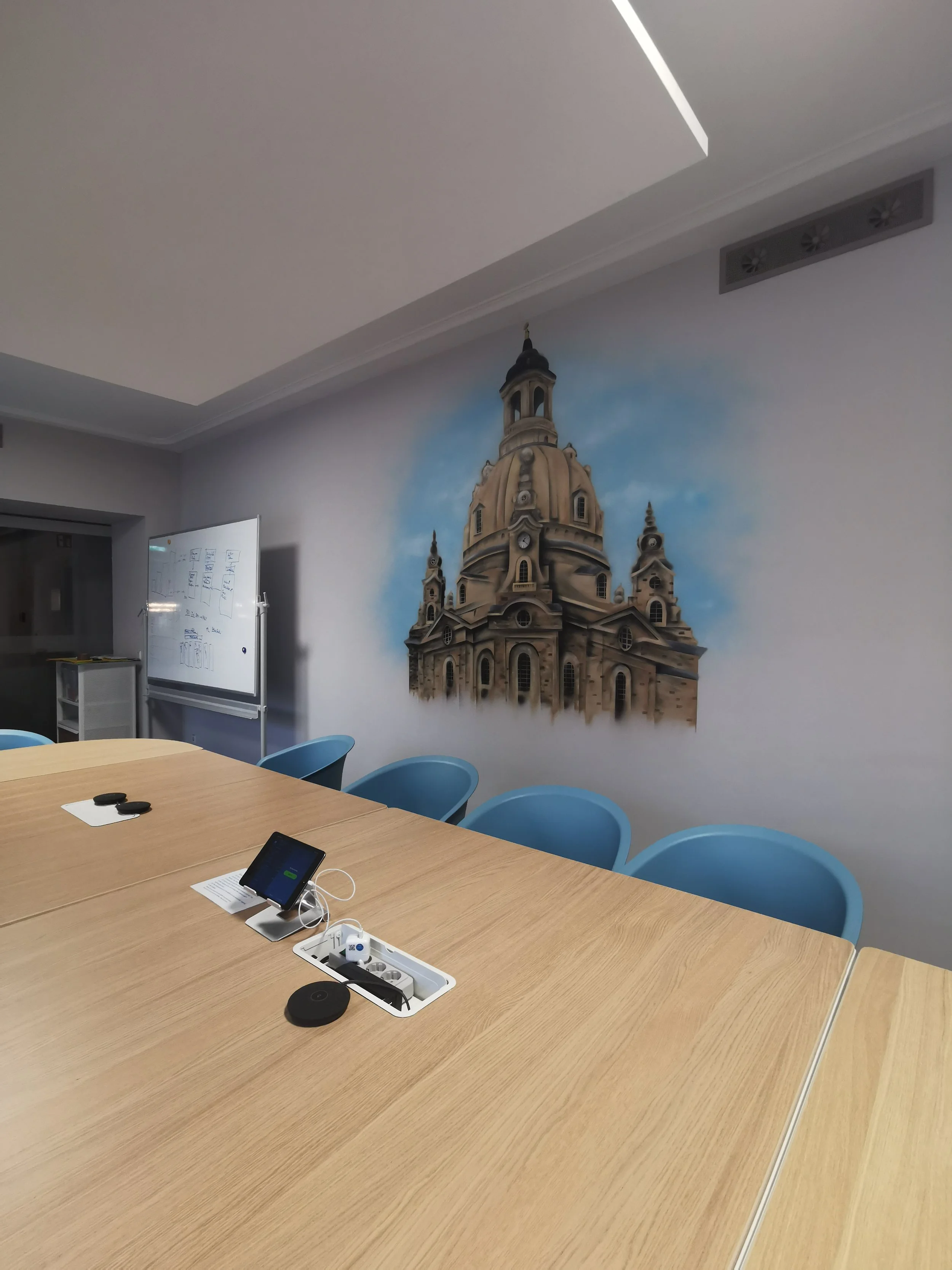 Ein Konferenzraum mit einem großen Tisch, blauen Stühlen, einem Whiteboard mit Notizen, einem Smartphone, einer Power-Strip mit Kabeln und einem Wandbild eines barocken Kirchturms an der Wand.