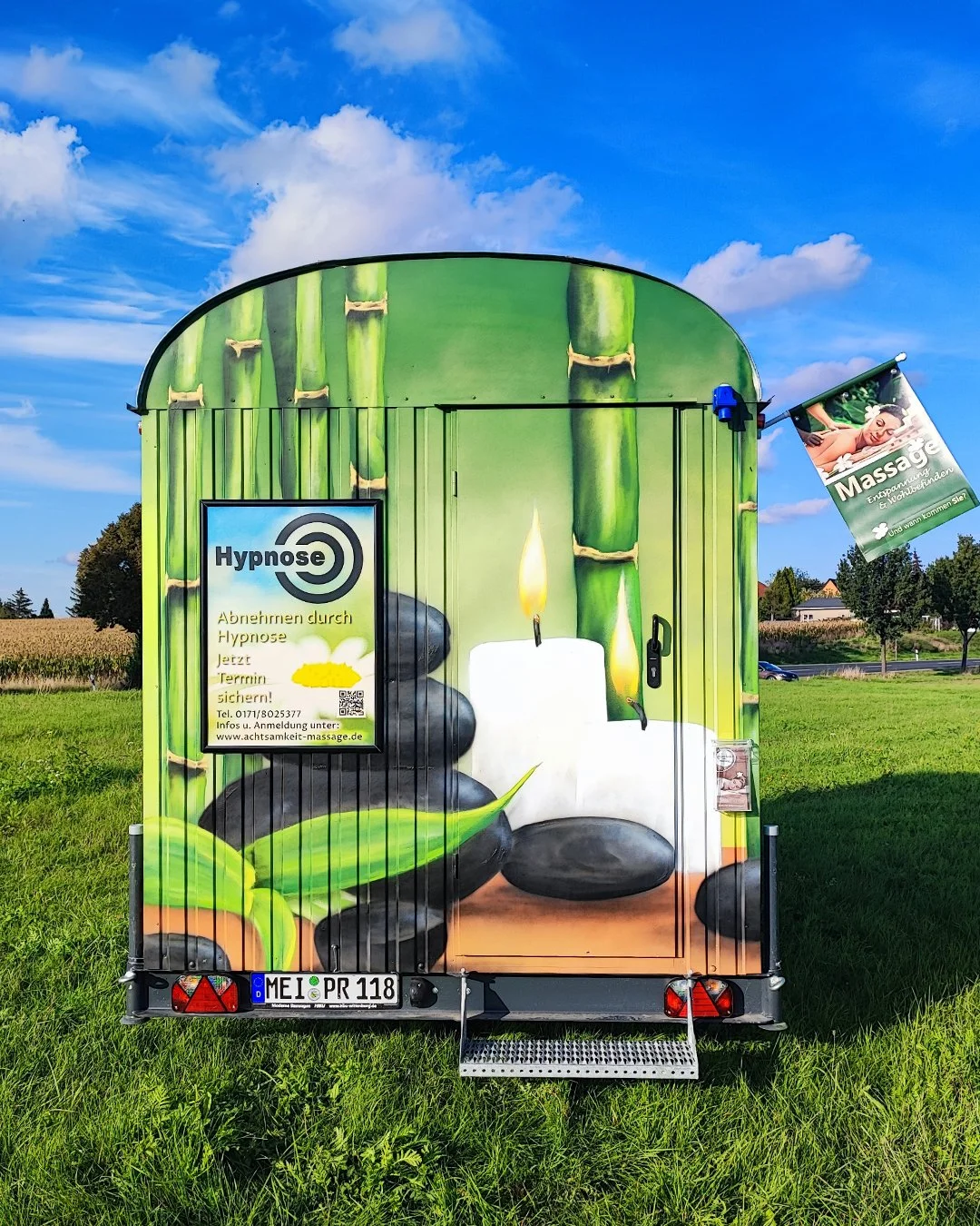 Mobile Massagewagen mit grünen Bambus- und Steinen im Design, Werbung für Hypnose und Massage, auf grünem Feld bei blauem Himmel.