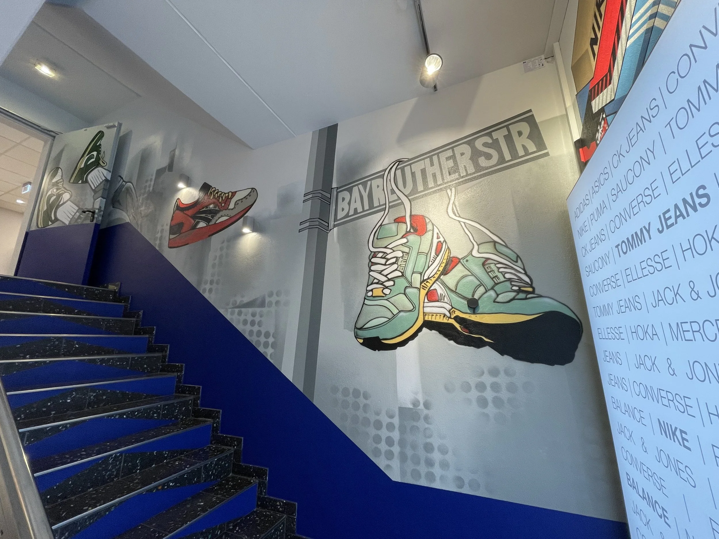 Graffiti Kunst an einer Treppenaußenwand, das Schuhe und eine Straßenbahnlinie namens 'Bayruther Str' zeigt.