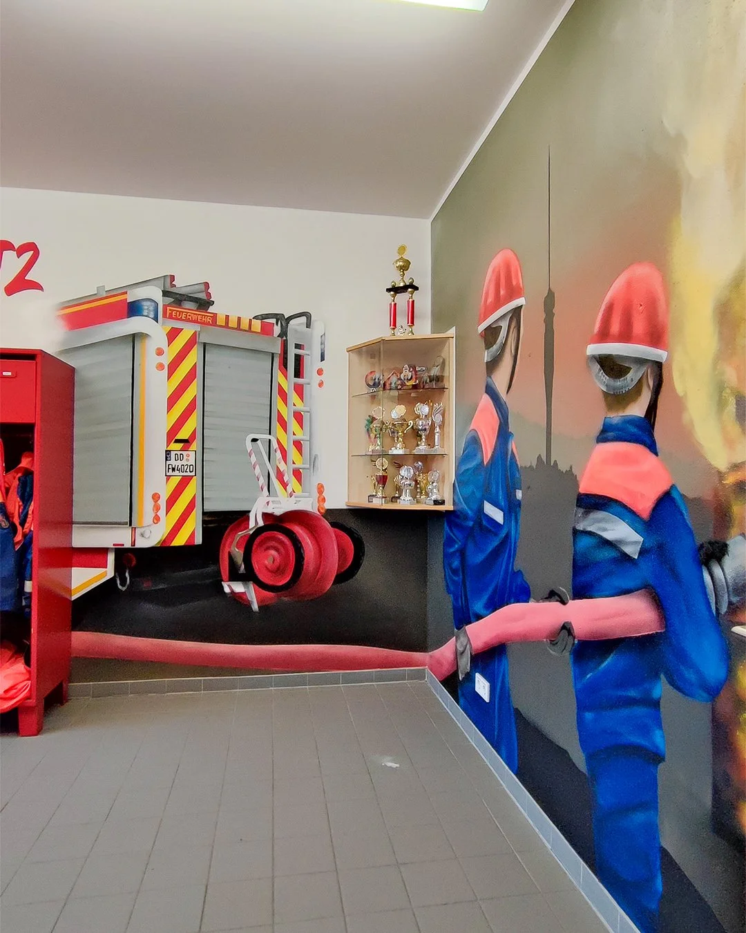 Mural mit Feuerwehrleuten in Uniformen und Helmen, Feuerwehrauto und Trophäen im Hintergrund.