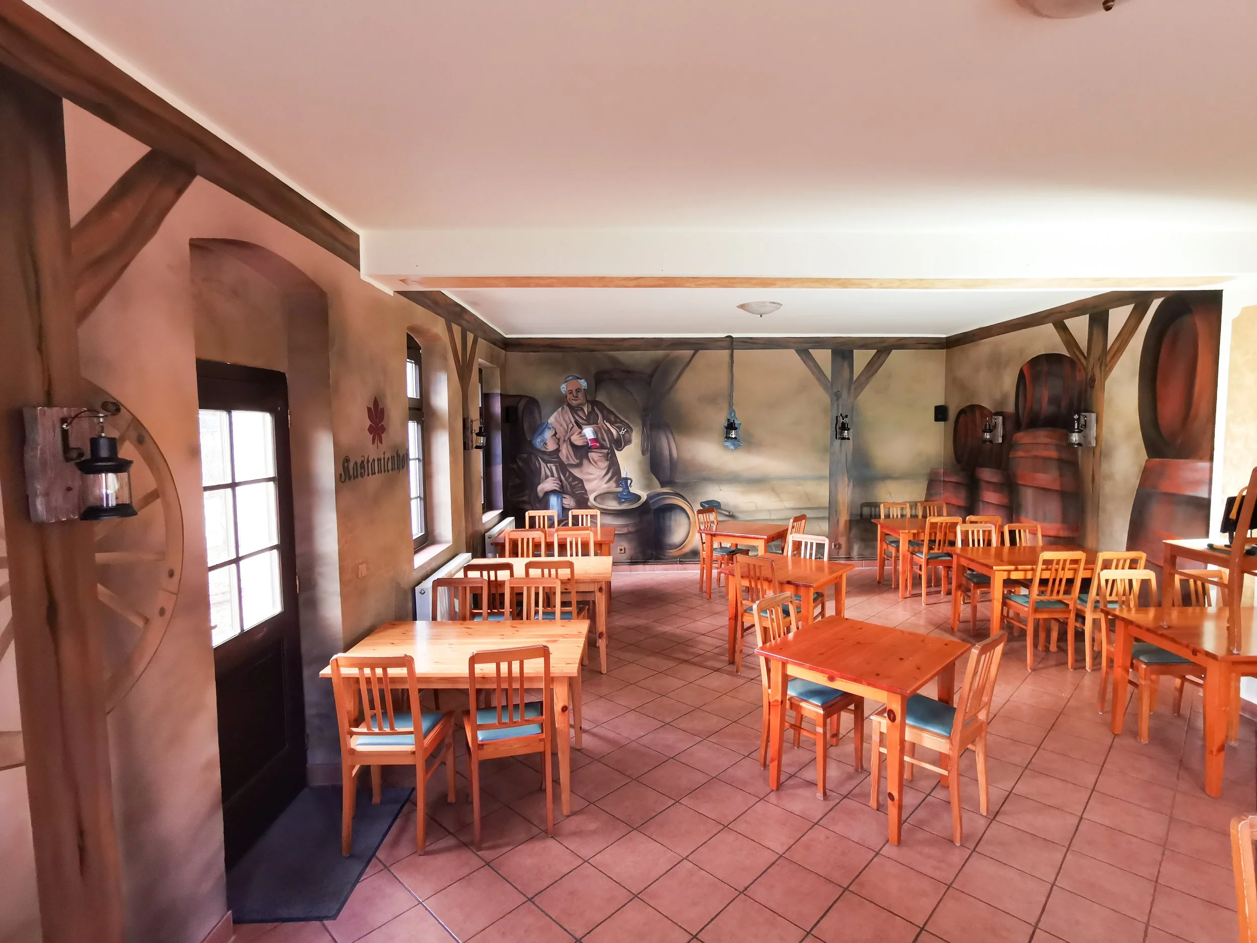 Innenansicht eines rustikalen Restaurants mit Holzstühlen und -tischen, Wandmalereien von Weinfässern und einer Szene mit einer Person, die Wein in einem Keller hält.