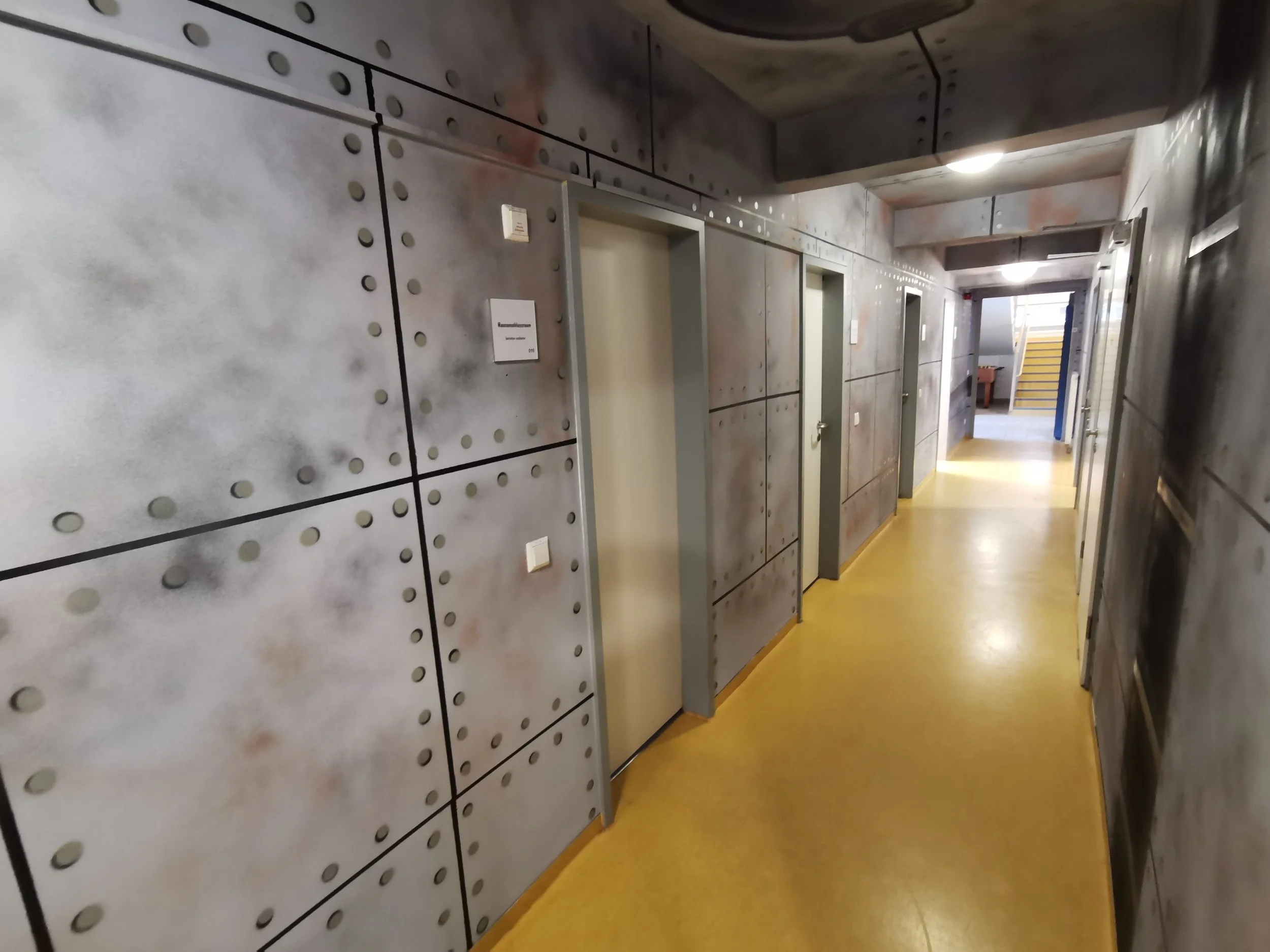 Ein Flur mit grauen Wänden mit schwarzen Linien und runden Mustern, gelber Boden, mehrere Aufzüge und Türen, Treppe im Hintergrund.