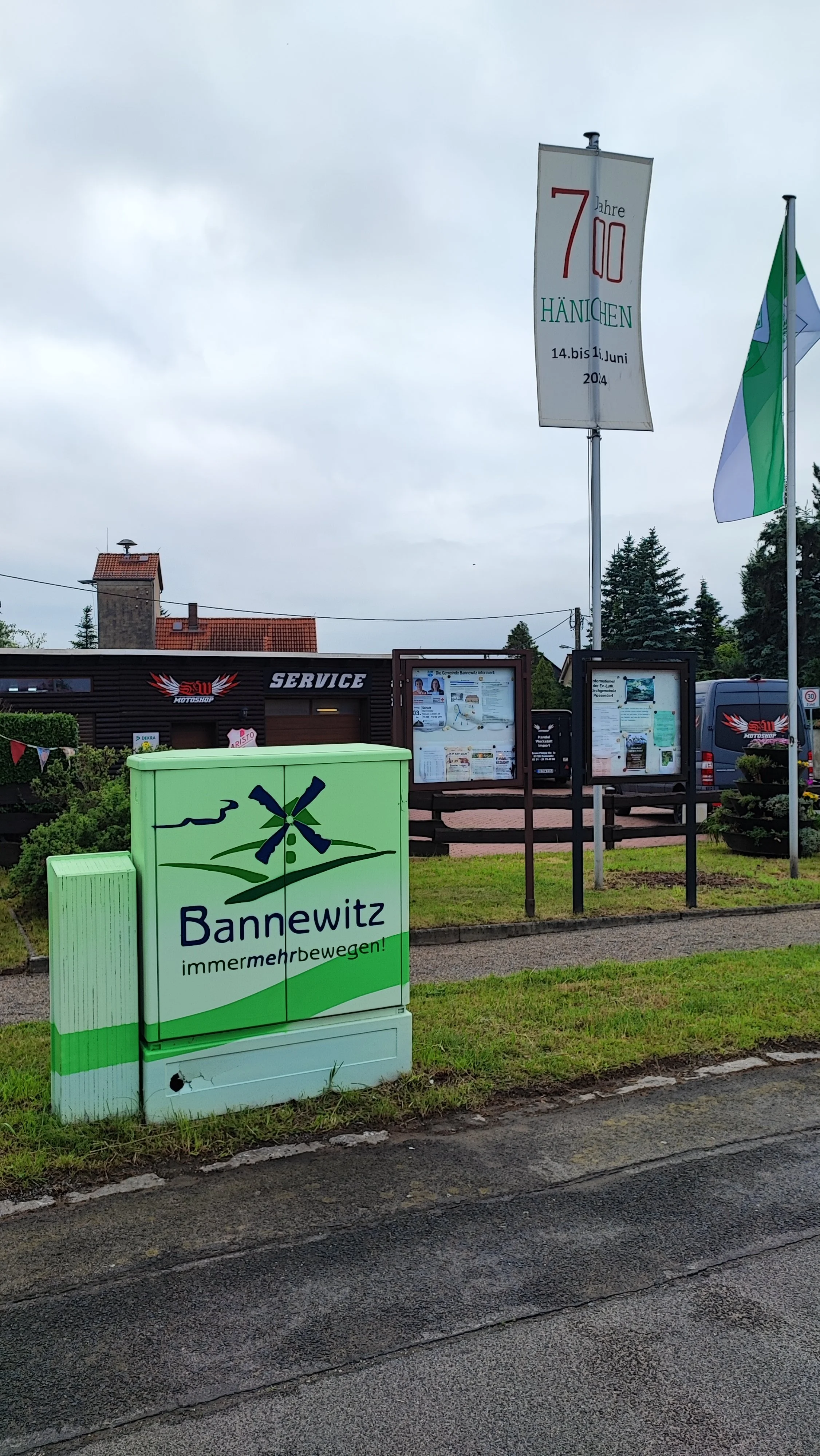 Ein Straßenbild mit einem grünlichen Behälter, der die Aufschrift 'Bannewitz immer mehr bewegen!' trägt, sowie ein Schild mit dem Text '70 Jahre Hänichen 14. bis 21. Juni 2014' und eine Flagge im Hintergrund.