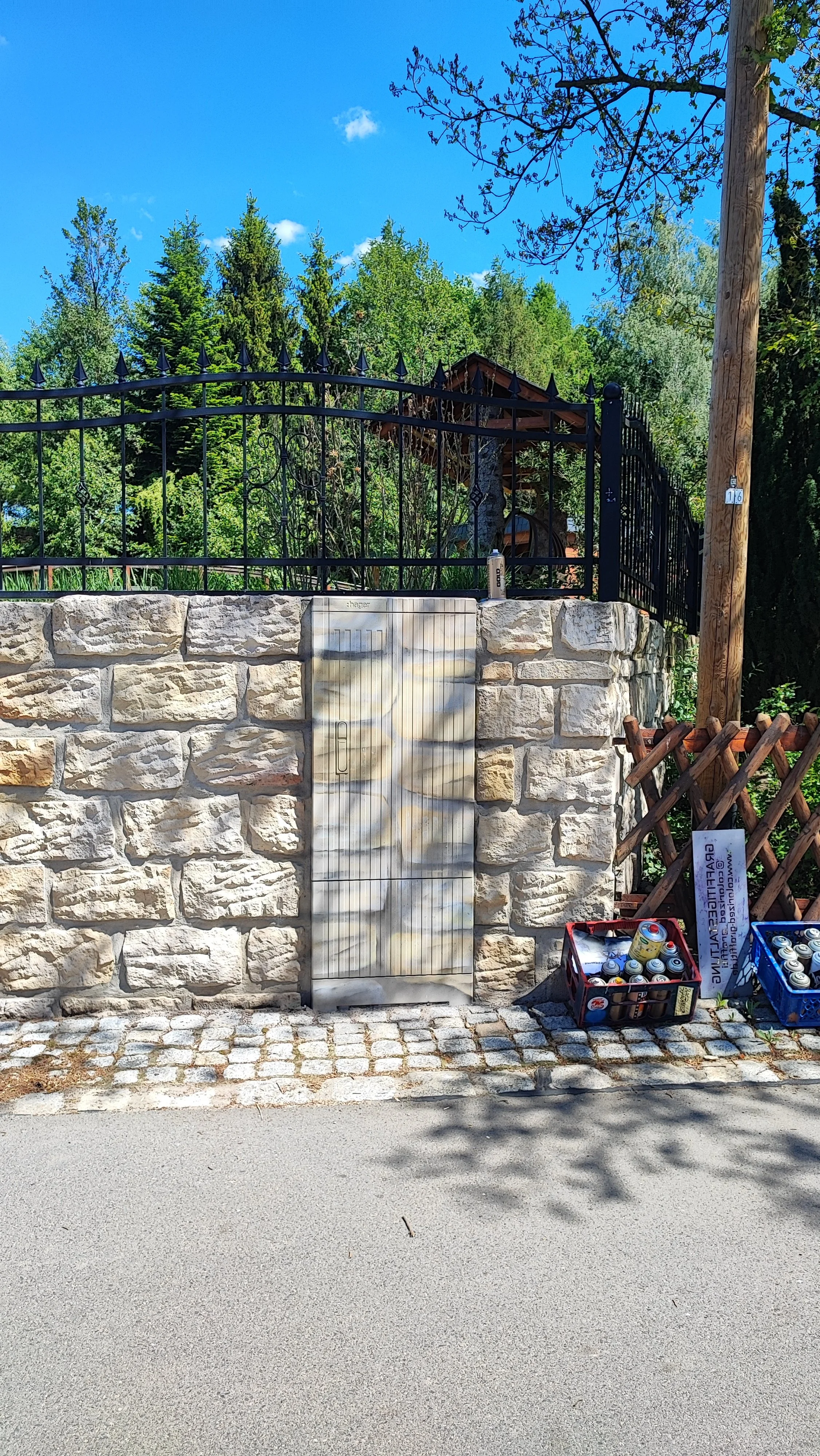 Ein Steinmauer mit einem Metallgitterzaun auf einem Höhenunterschied, daneben stehen zwei Kästen mit Getränkedosen. Es sind Bäume und ein blauer Himmel sichtbar.