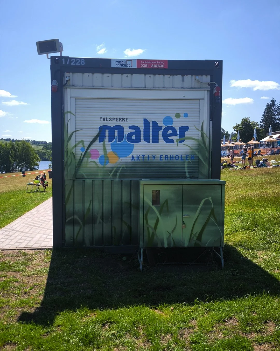Ein kleiner Kiosk mit einem gemalten Gras-Design auf der Tür, befindet sich in einer parkähnlichen Umgebung mit Wasser im Hintergrund. Das Schild auf dem Kiosk liest 'Talsperre Malter aktiv erholen'.