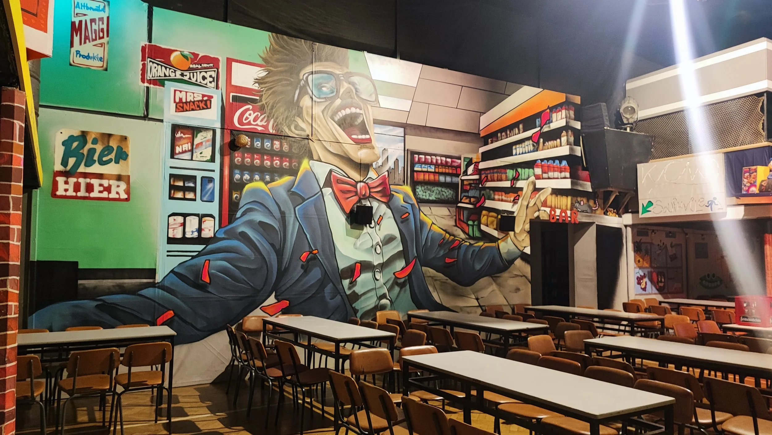 Street art Murale mit lachendem Mann in Anzug, der in einem Supermarkt steht, umgeben von Regalen und Schildern, mit Tischen und Stühlen im Vordergrund.