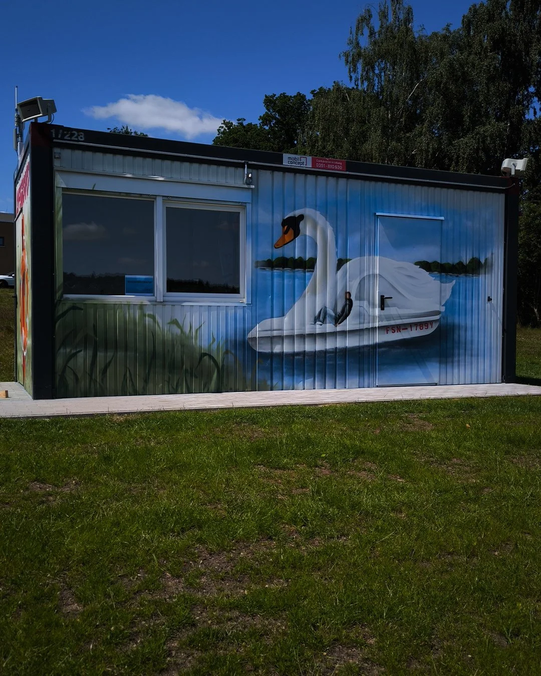 Malerische Wandmalerei auf einer kleinen Hütte: Ein Schwan mit einem kleinen Kind auf seinem Rücken, im Wasser mit einer natürlichen Uferlandschaft im Hintergrund.