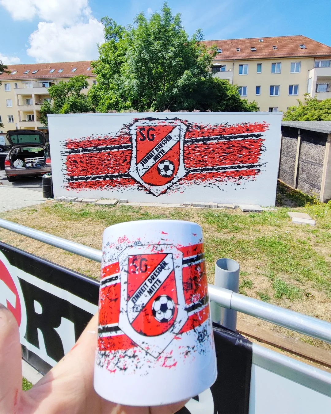Ein Graffiti an einer weißen Wand zeigt das Logo eines Fußballvereins mit dem Text 'SG' und 'EINHEIT DRESDEN MITTE', darunter ein Fußball. Im Vordergrund hält jemand eine Tasse mit dem gleichen Logo.
