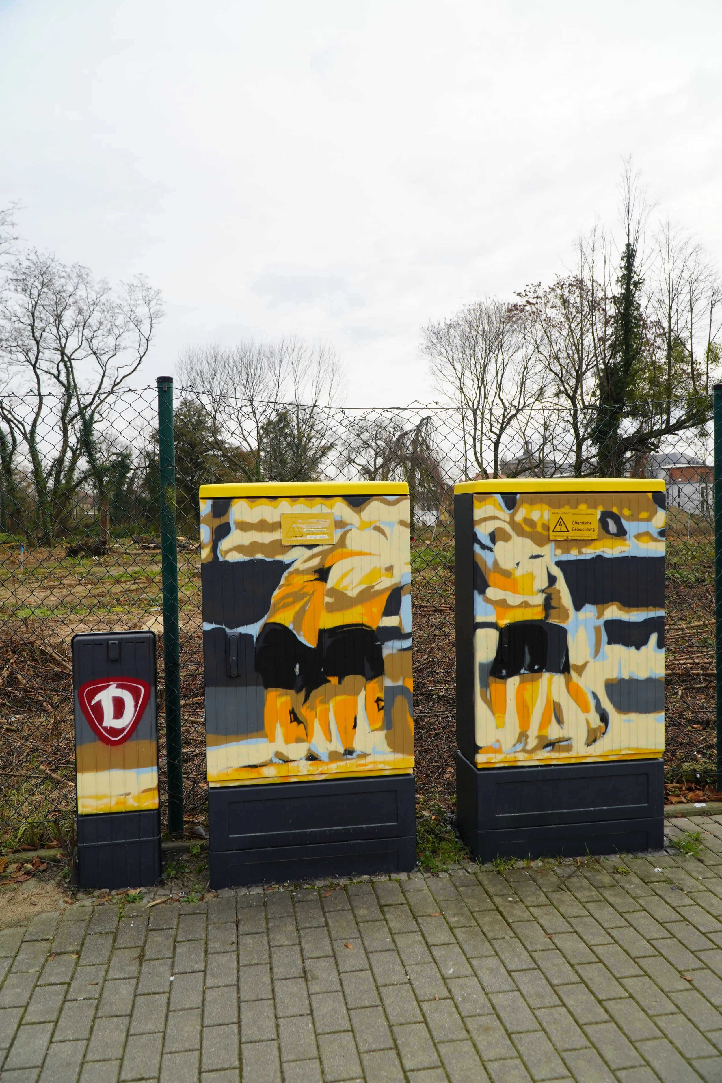 Graffiti bemalte Stromkästen, die Kinder beim Spielen darstellen, vor einem Zaun und Bäumen auf einem gepflasterten Boden.